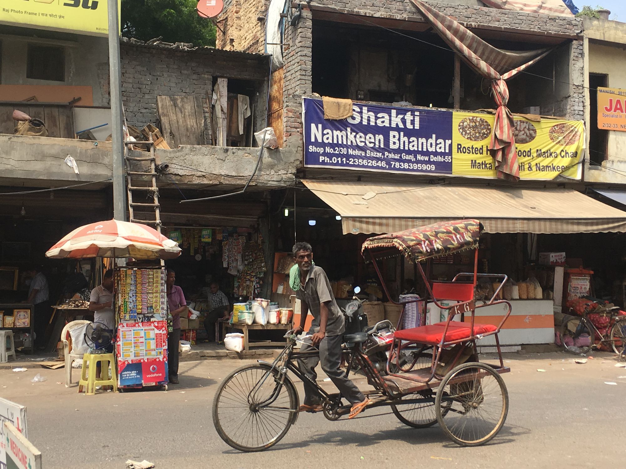 A man rides a bicycle-powered passenger cart past storefronts and umbrellas on a street in New Delhi