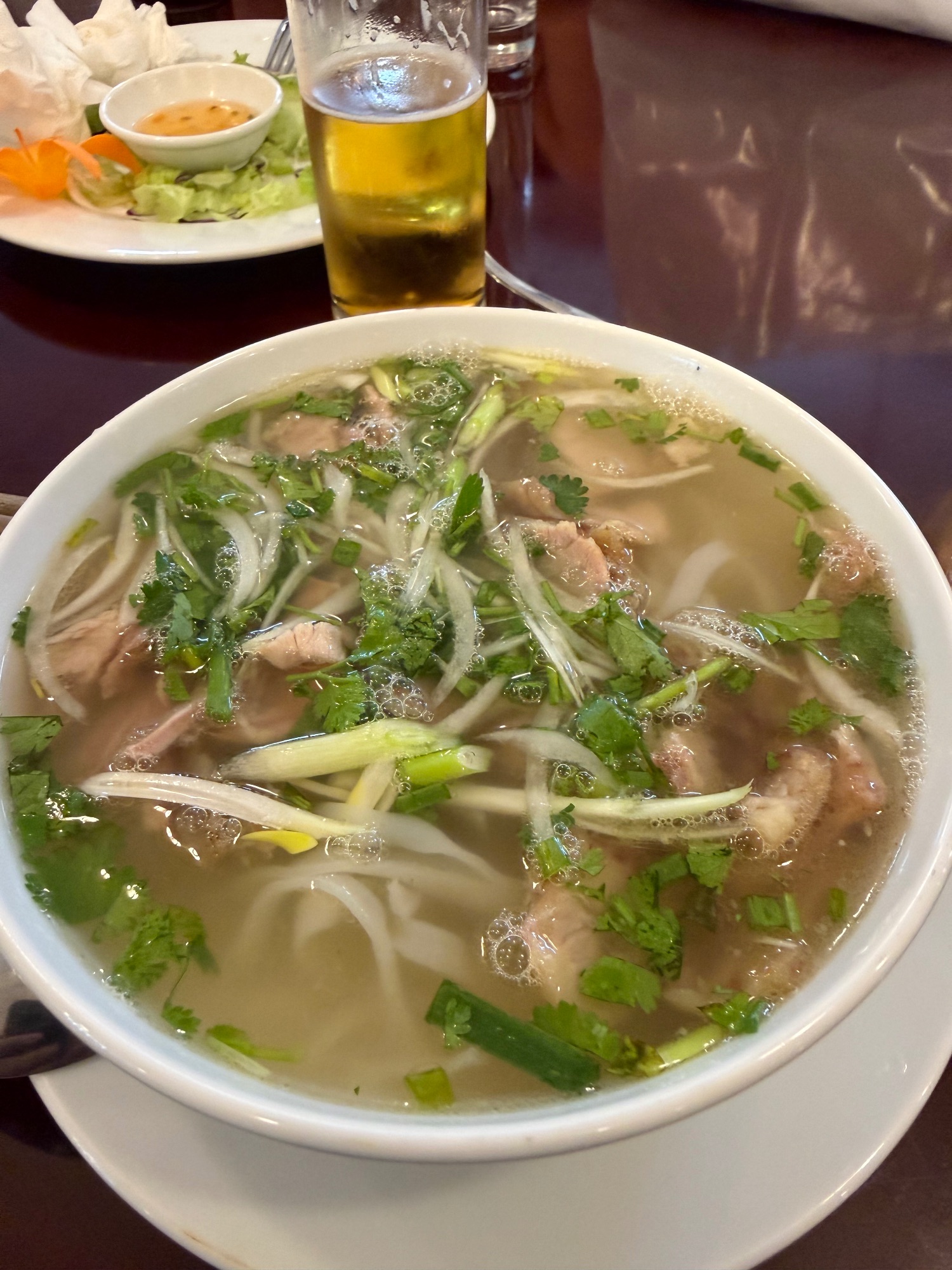 Bowl of pho with beef, noodles, and fresh herbs in clear broth, served with a glass of iced tea