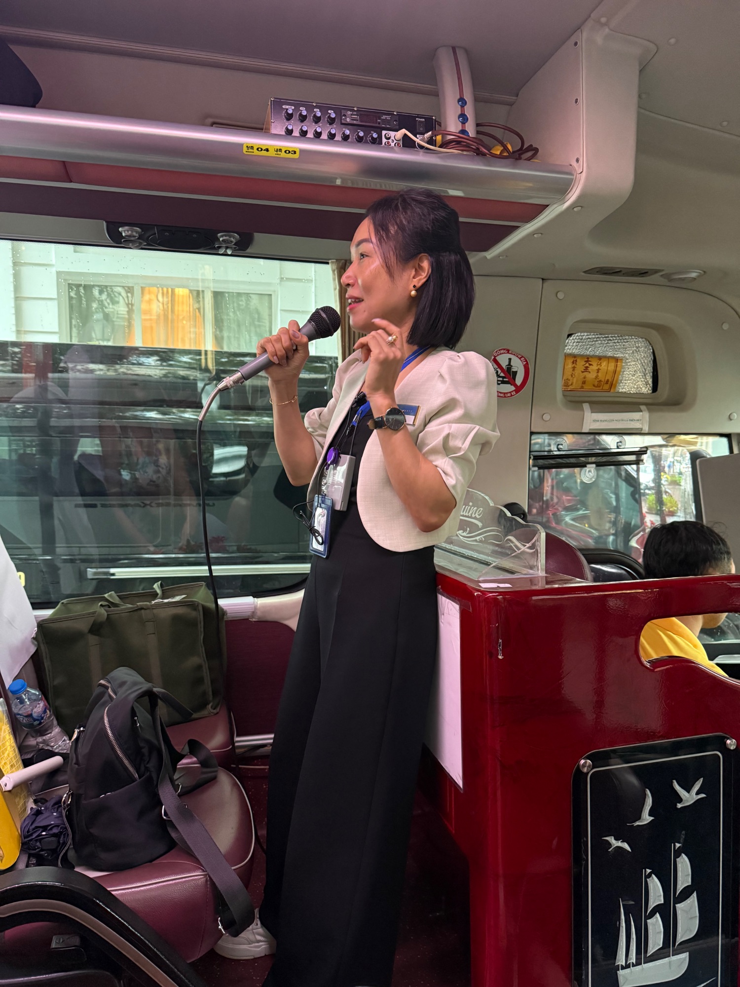 Woman speaking into a microphone on a tour bus, wearing professional attire and ID badge