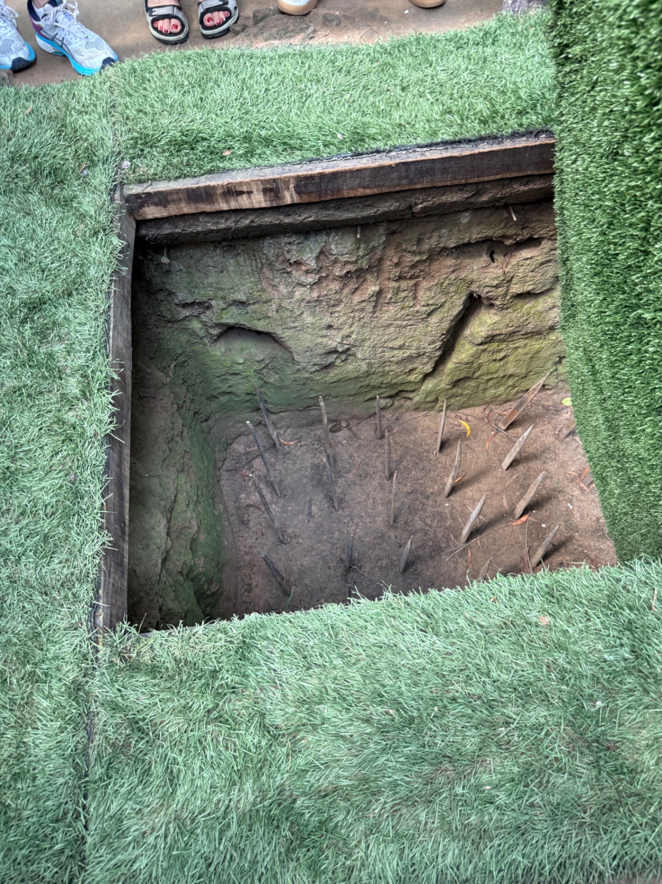 Footwear and legs visible above a rectangular pit with layered soil and archaeological markings