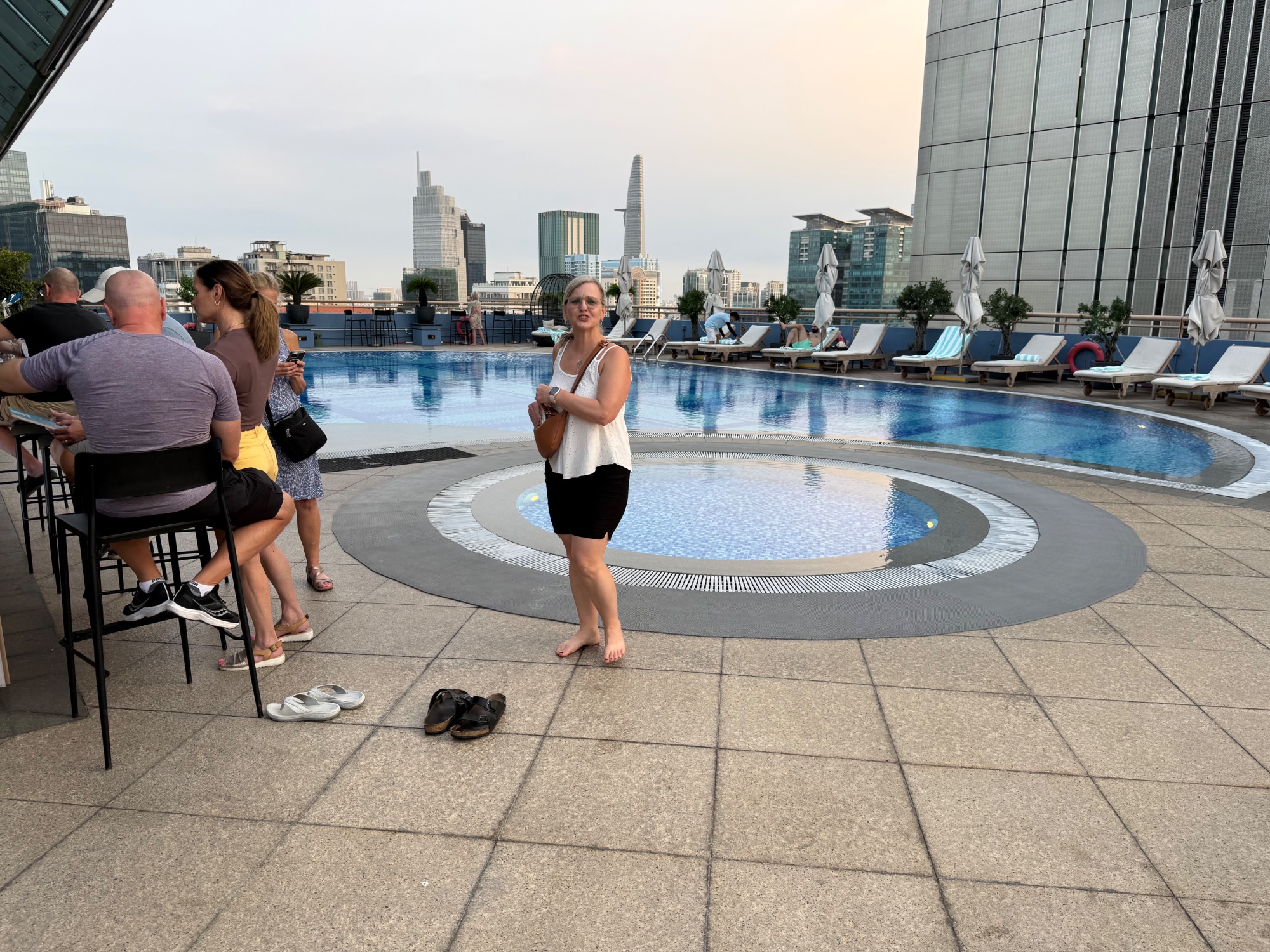 Woman in white tank top standing barefoot by a rooftop pool overlooking San Francisco's skyline