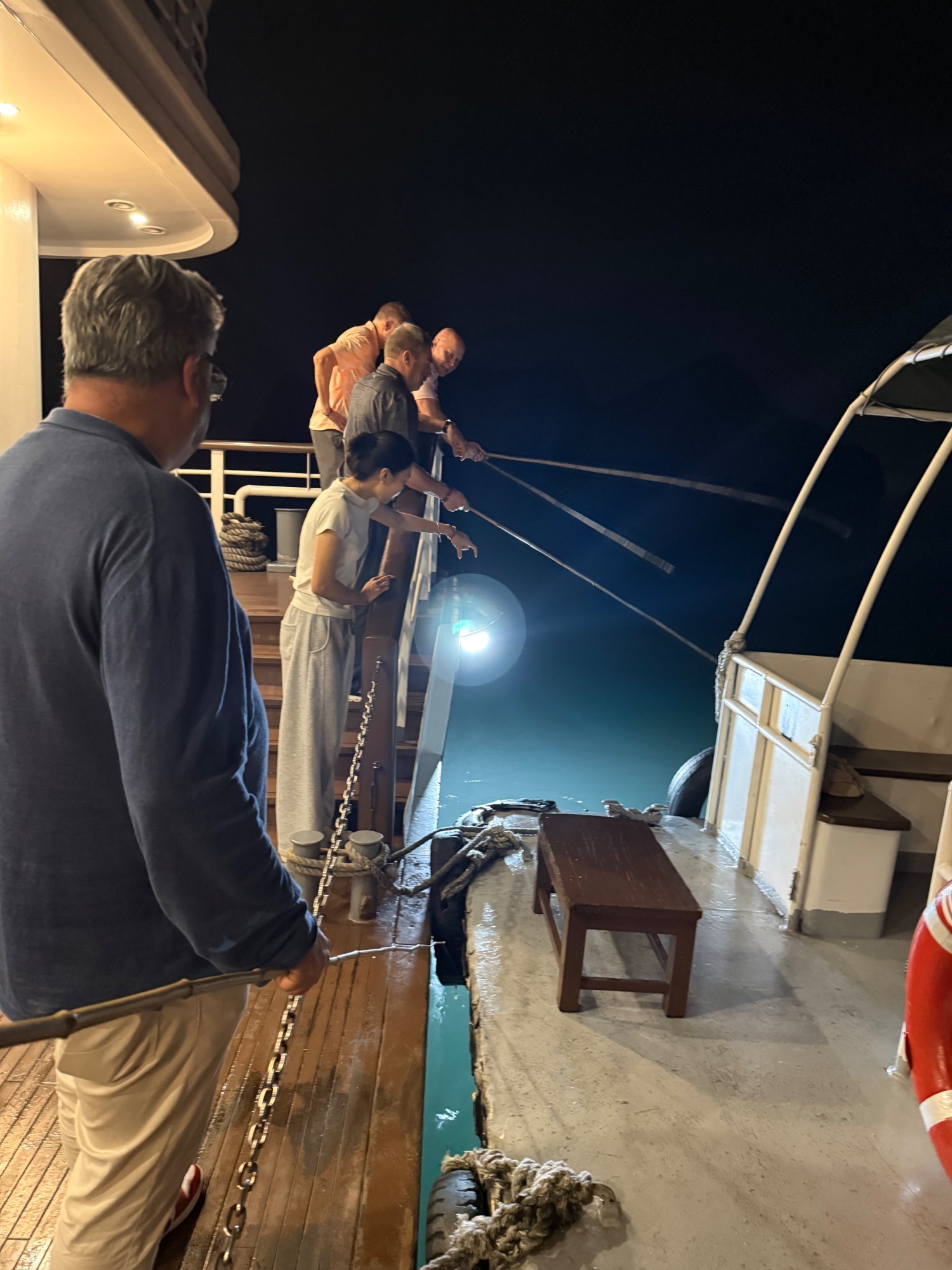 Group of tourists on a boat deck at night, some leaning over the railing to view the water below