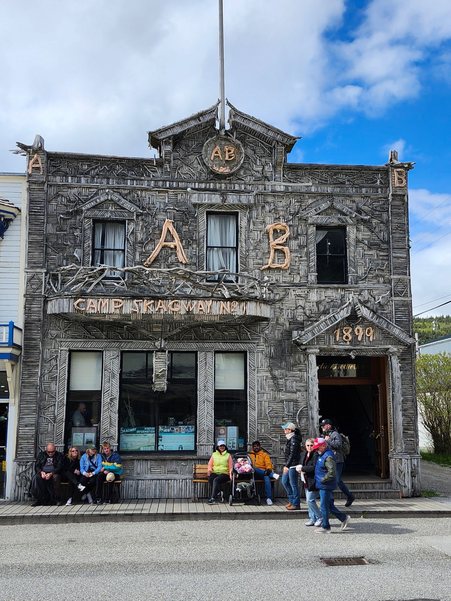 Ornately carved wooden building with 'Camp Skagway' sign and large numbers, tourists sitting on bench outside, dated 1898