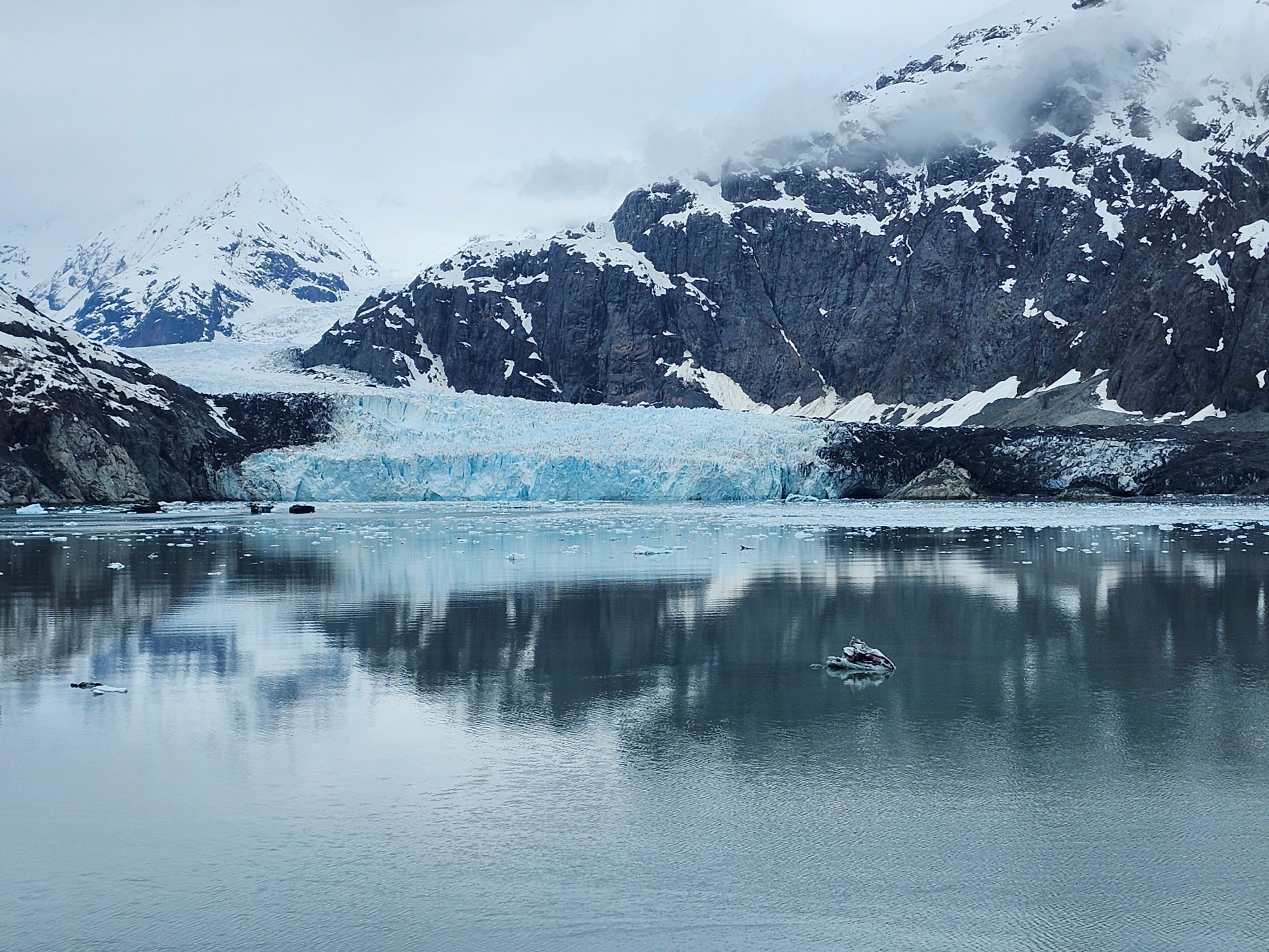 Bright blue glacier meeting calm water surrounded by snow-covered rocky peaks and mountains in Alaska