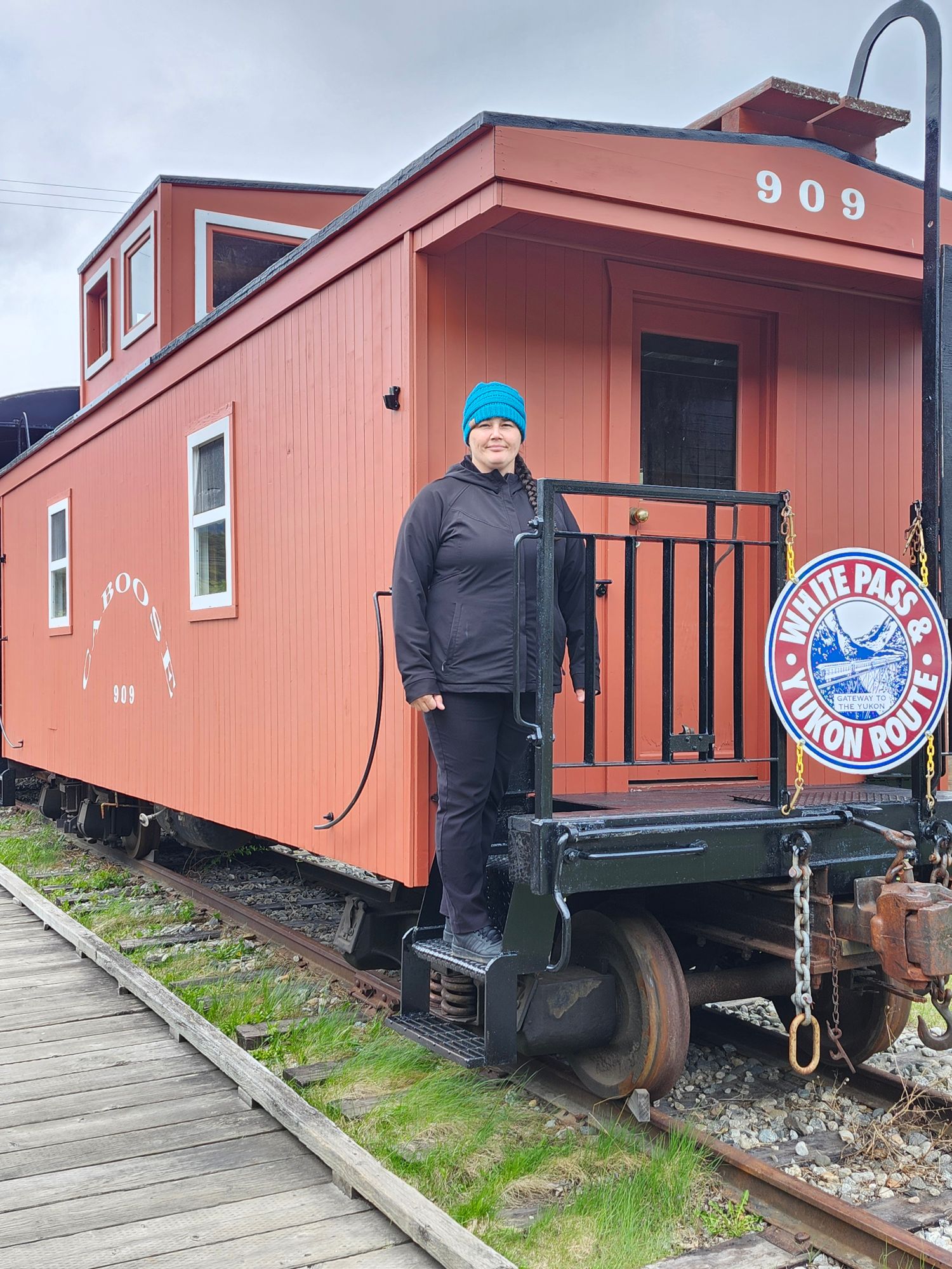 Echo Brocato standing on platform of red caboose numbered 909 with White Pass & Yukon Route railroad sign