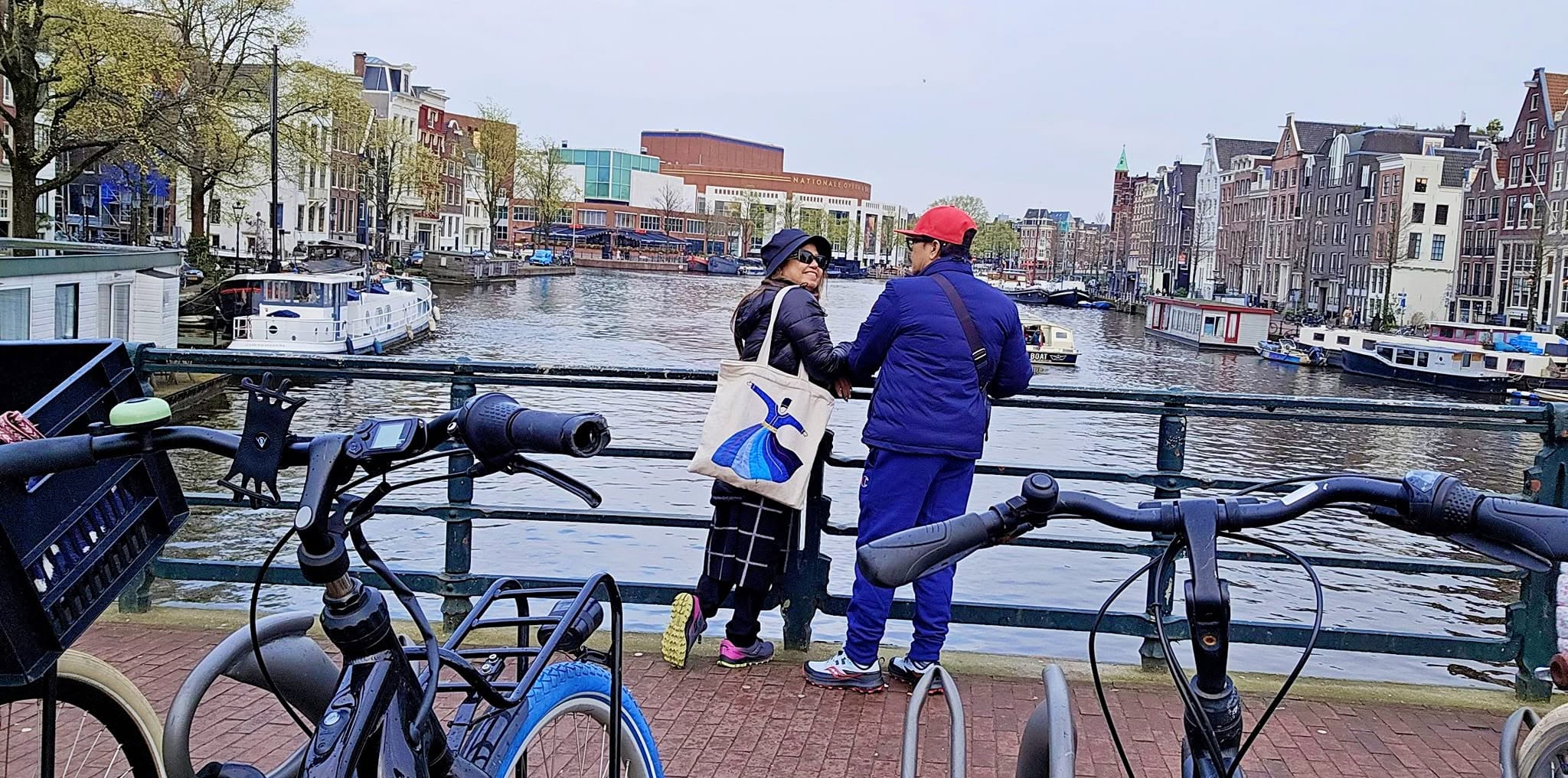 Two cyclists in blue outfits standing with bikes on Amsterdam canal waterfront with historic buildings