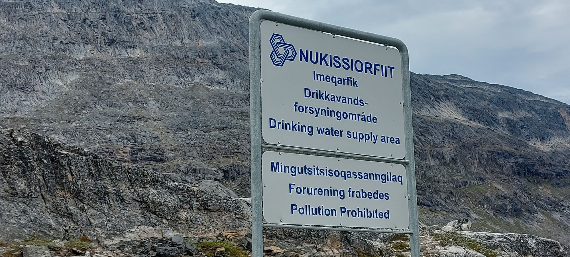 White sign marking Nukissiorfiit drinking water supply area with Greenlandic text on rocky, barren mountainside