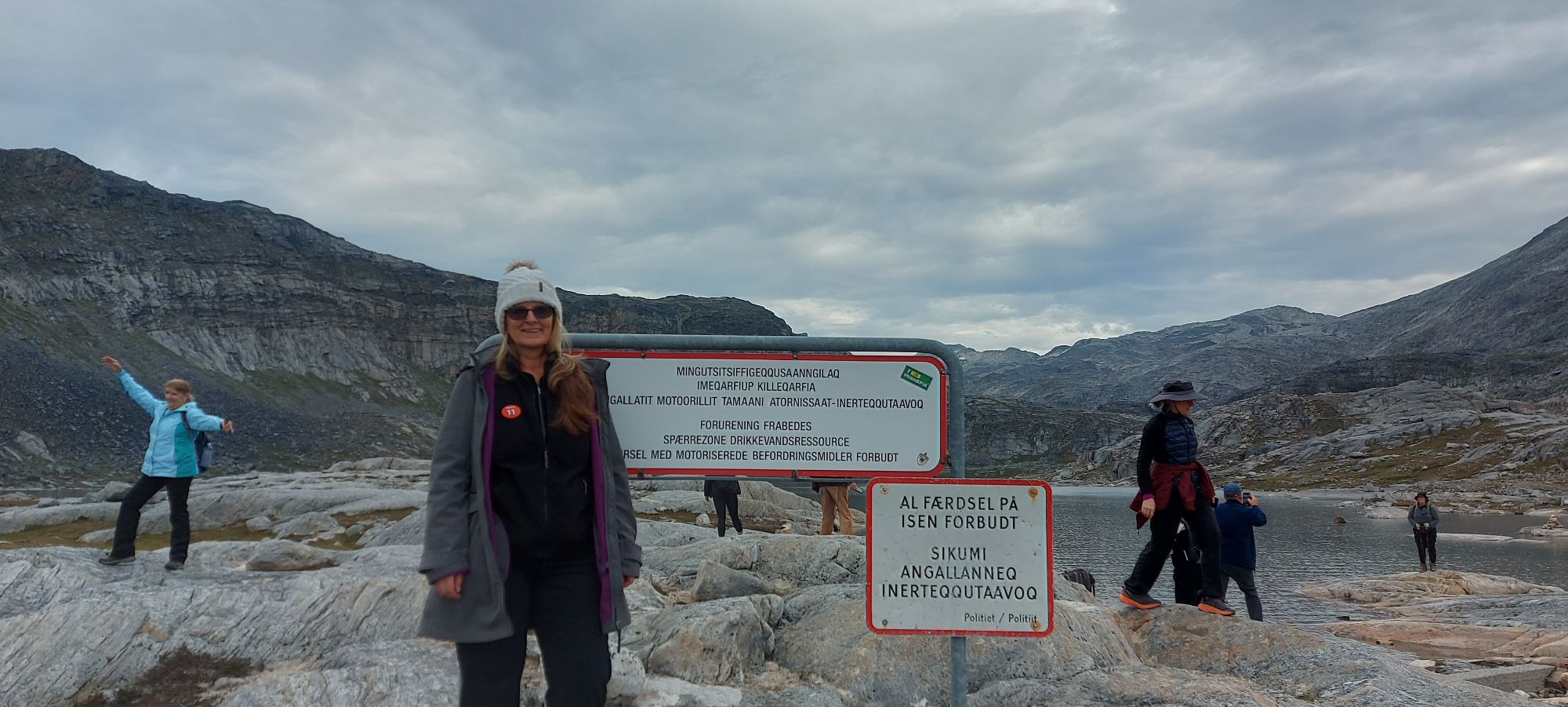 Woman in white hat standing by informational sign at glacial lake with rocky mountains and hikers in background
