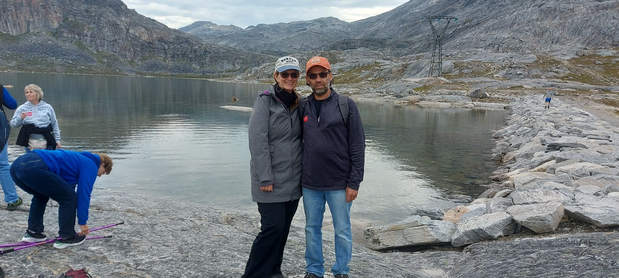 Couple posing by red warning sign at pristine glacial lake surrounded by steep rocky peaks and tundra landscape
