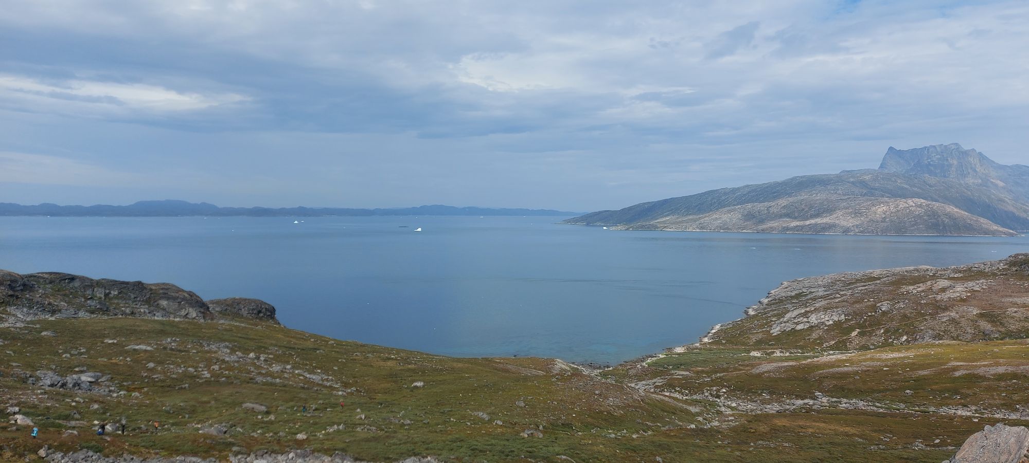 Iceberg floating in calm fjord waters with mountains and distant islands visible across the bay