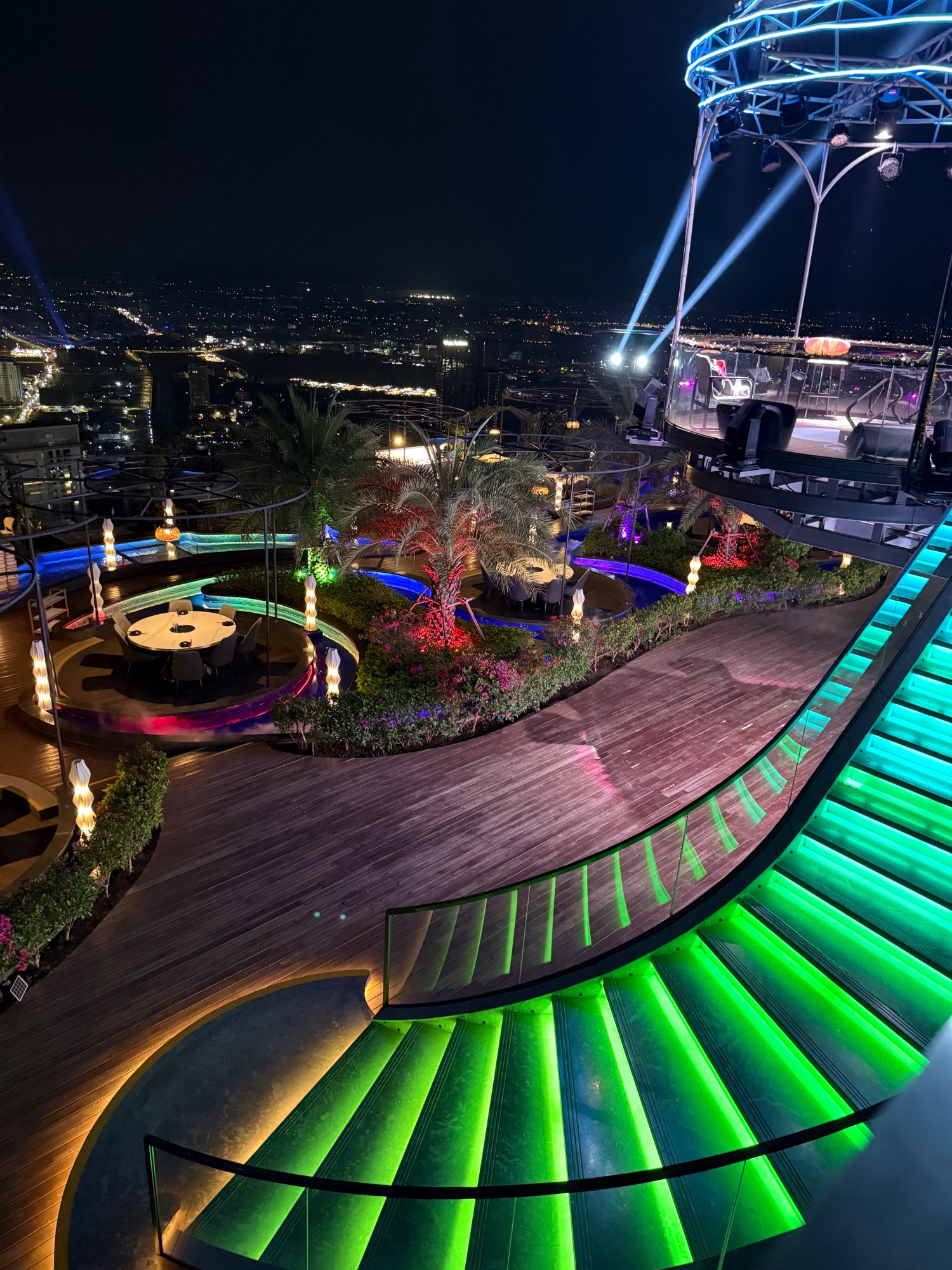 Illuminated rooftop garden and bar area at night with colorful LED lighting, decorative plants, and a stage with performance equipment
