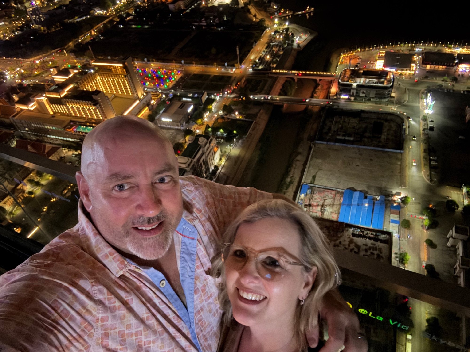 Man and woman taking a selfie on a high-rise balcony overlooking a brightly lit city at night with visible waterfront