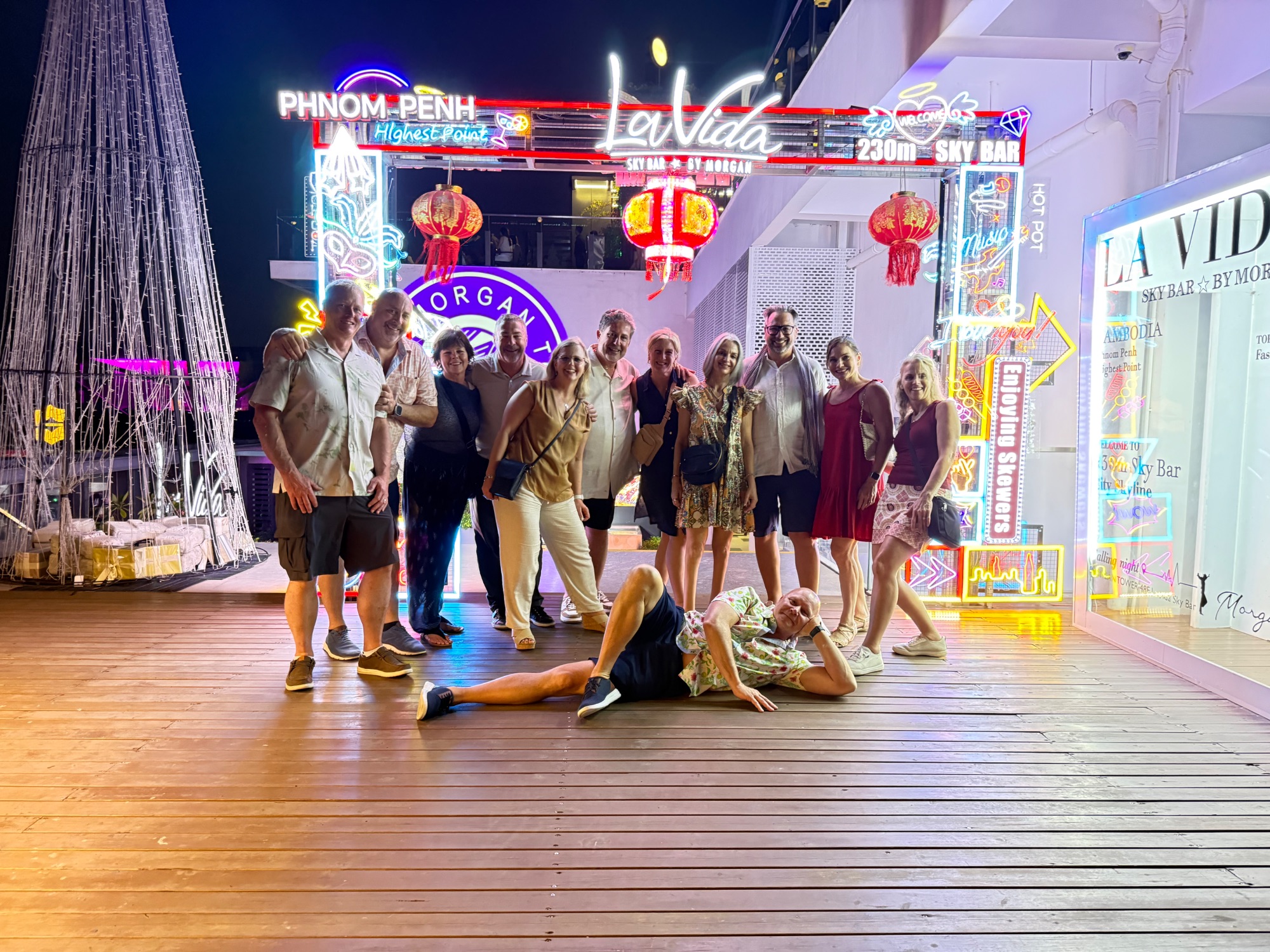 Large group of tourists posing outside a neon-lit Phnom Penh street market at night with colorful signage