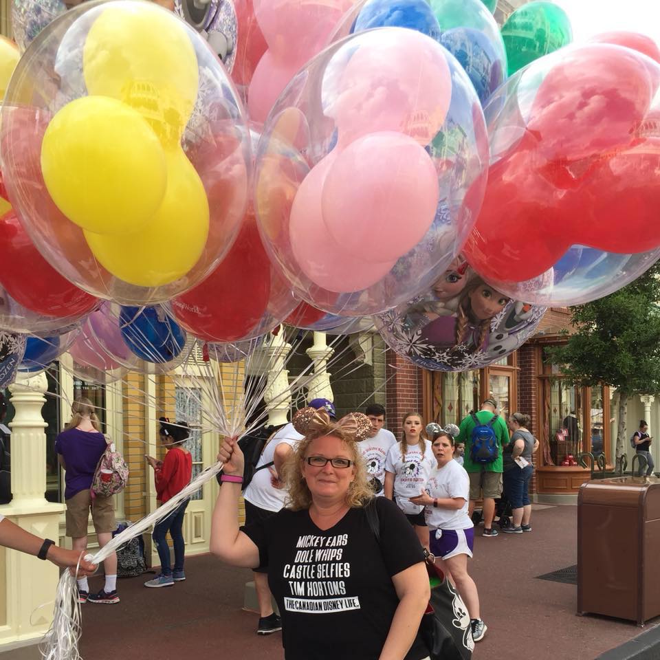 MaryBeth holding colorful character balloons on a Disney park street during daytime with crowds visible
