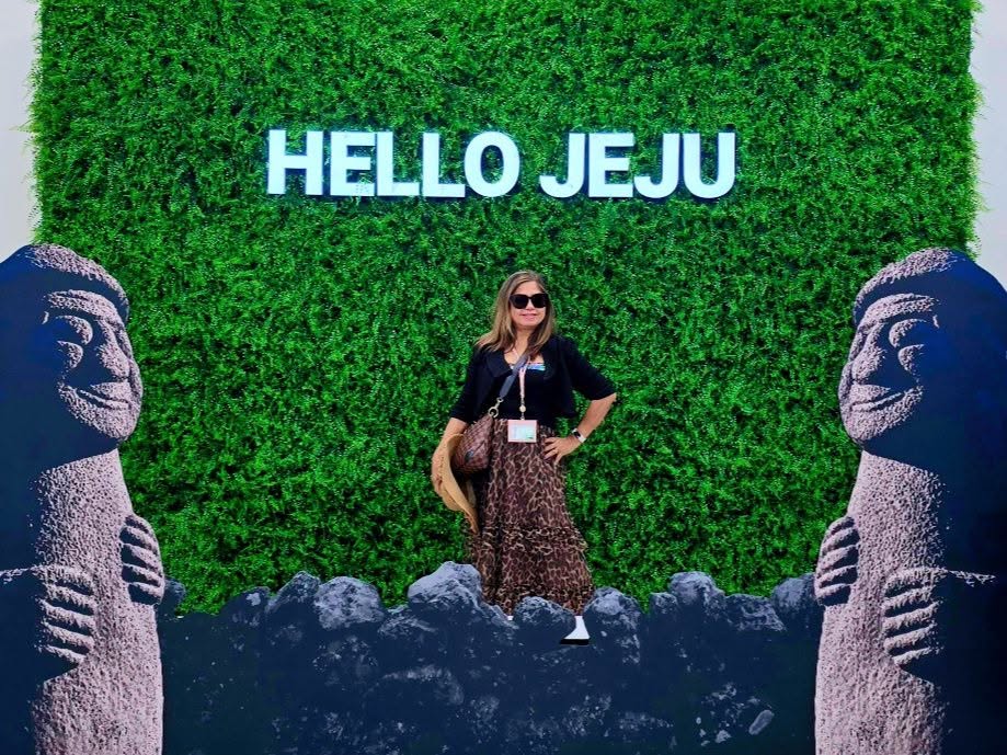 Woman standing at Hello Jeju sign with green moss wall and stone statues