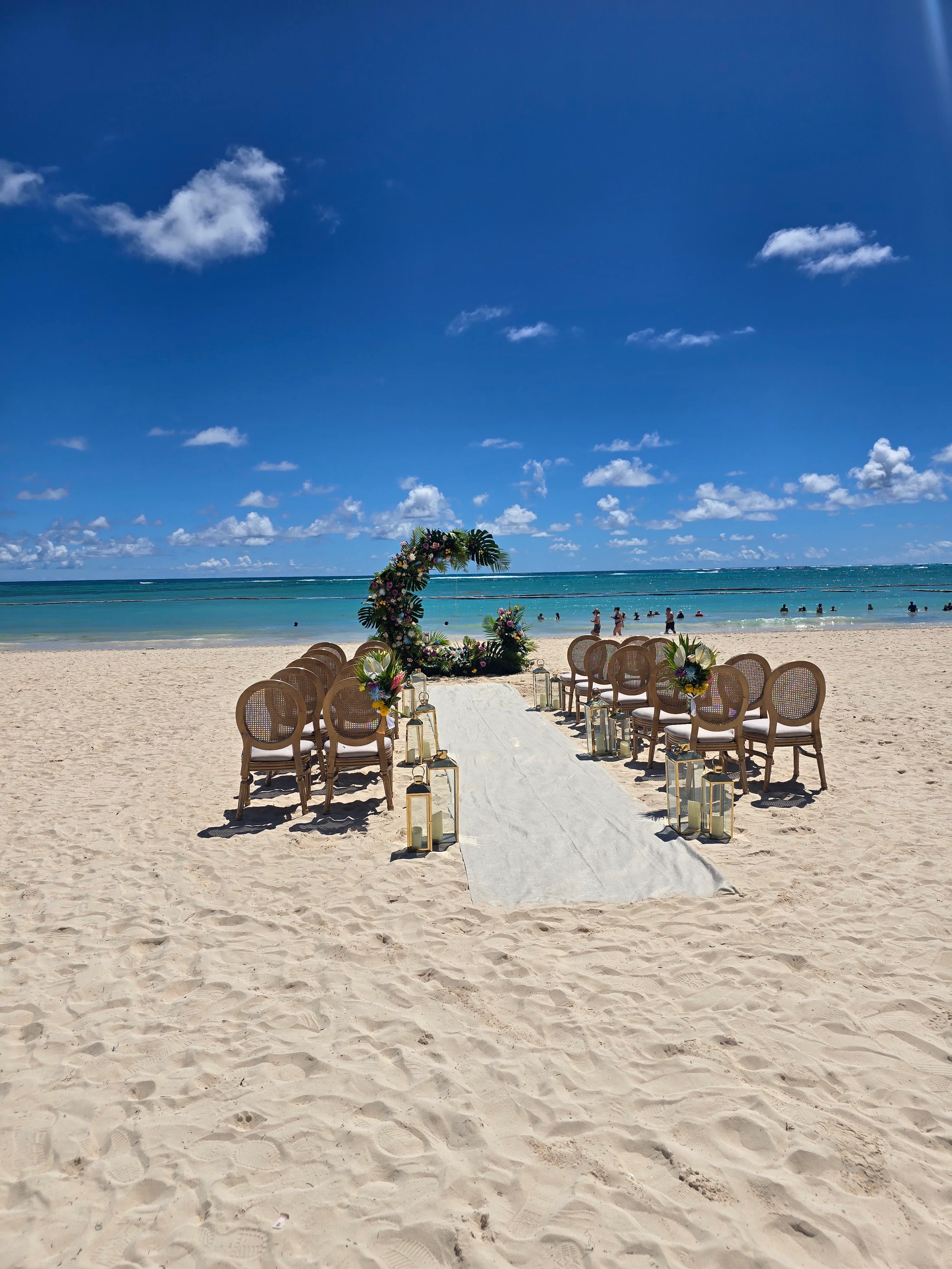 Beach wedding ceremony with rattan chairs arranged on sand, white aisle runner, floral arch, and swimmers in turquoise water