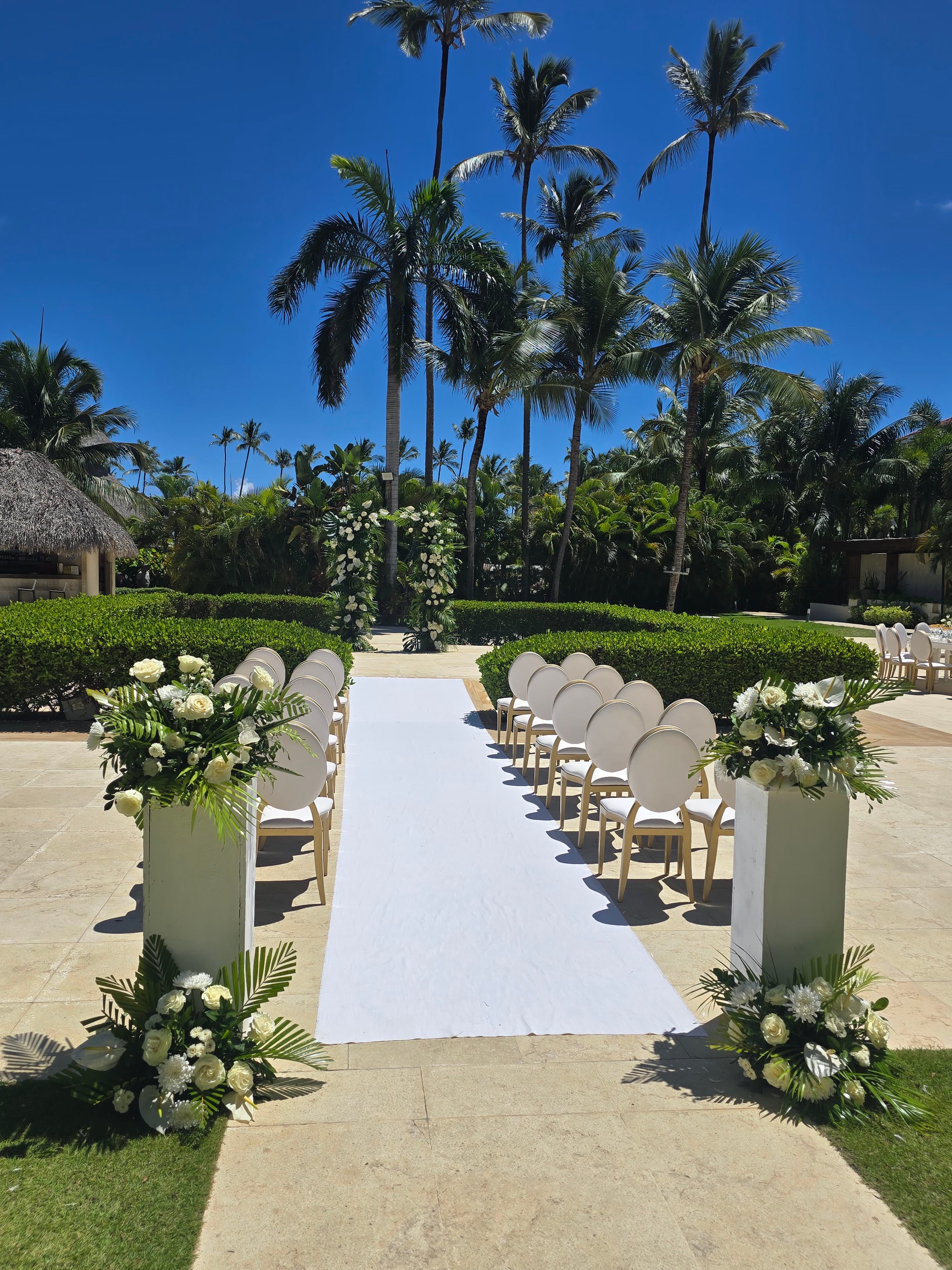 Outdoor wedding aisle on a paved courtyard lined with white chairs and white floral arrangements, palm trees and blue sky visible