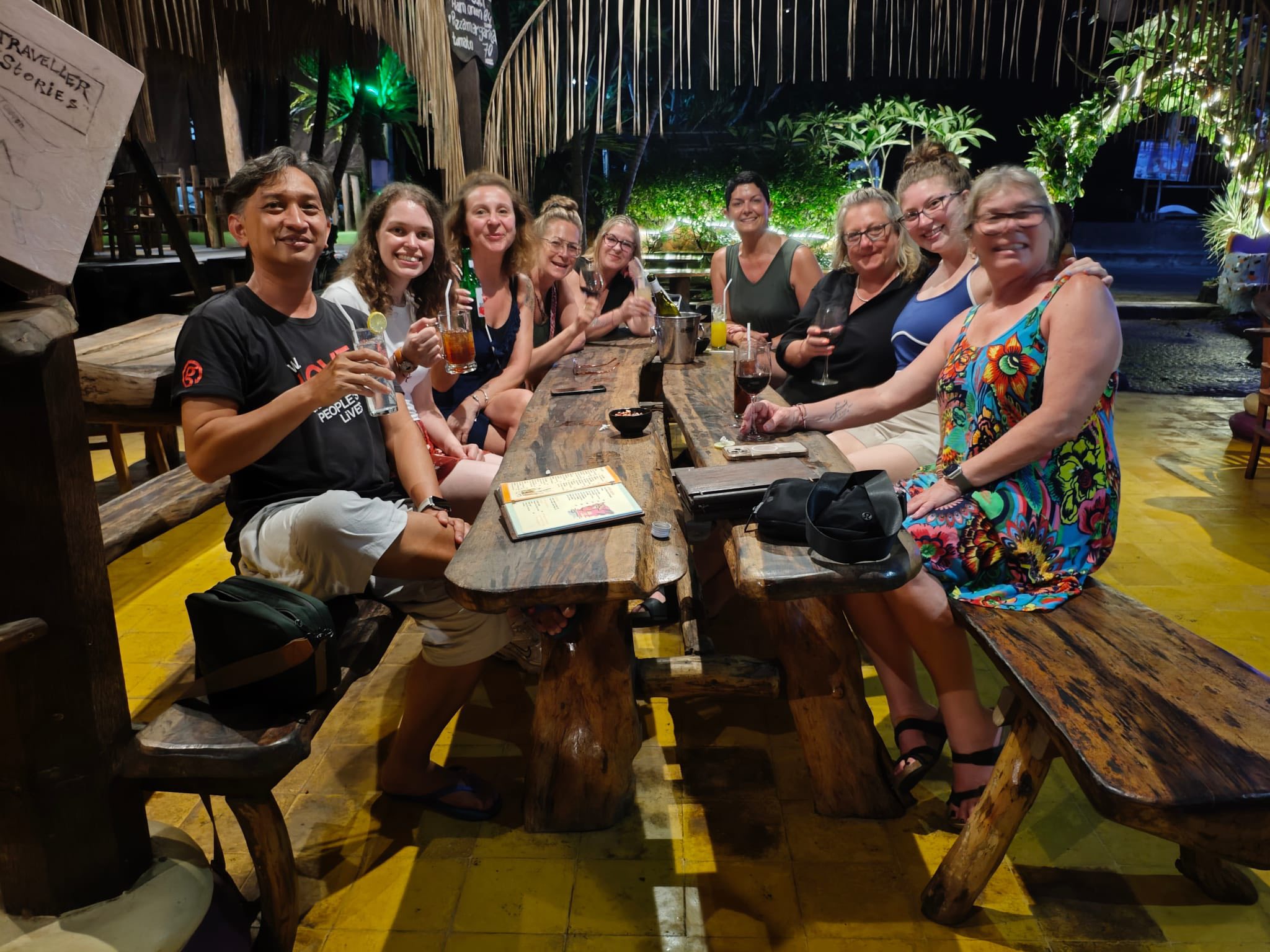 Diverse group of people seated at a long wooden table outdoors at night with drinks and smiling at camera