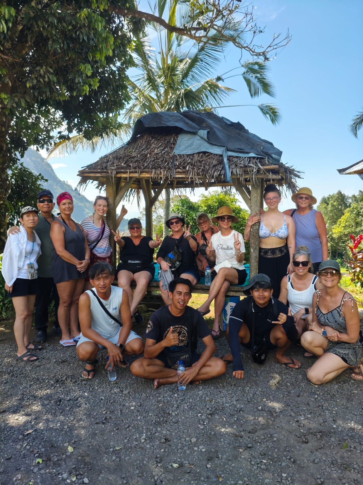 Group of tourists posing under a thatched-roof shelter with mountainous landscape visible in background