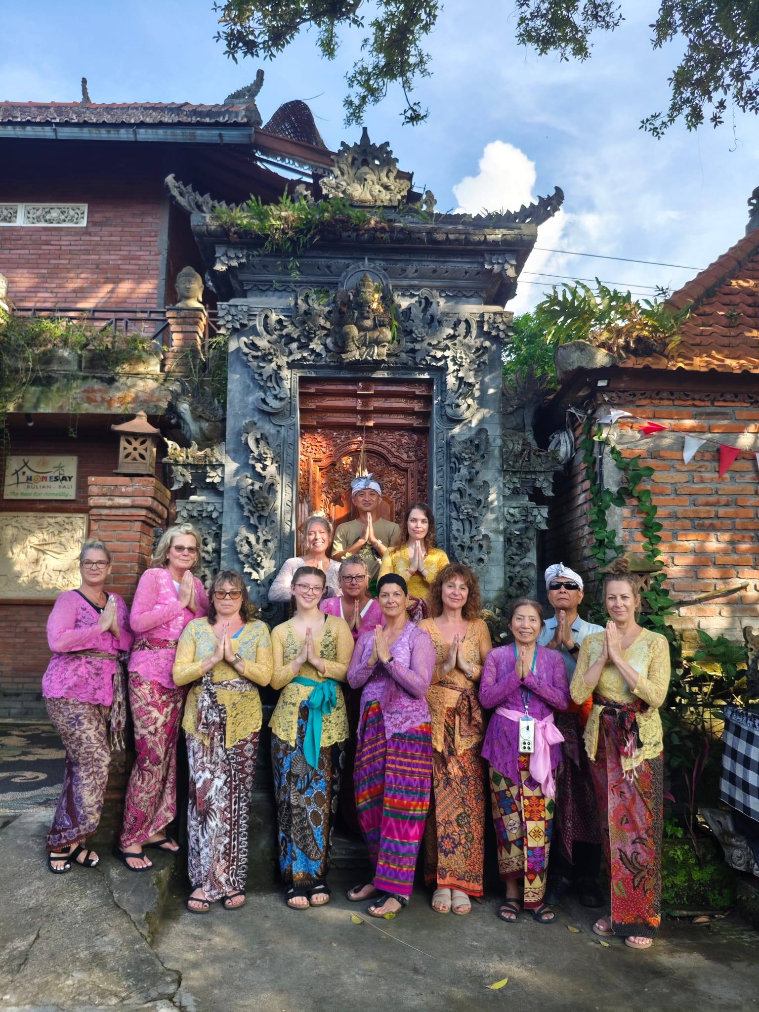 Group wearing traditional Balinese sarongs and sashes posing in prayer position outside an ornate temple entrance