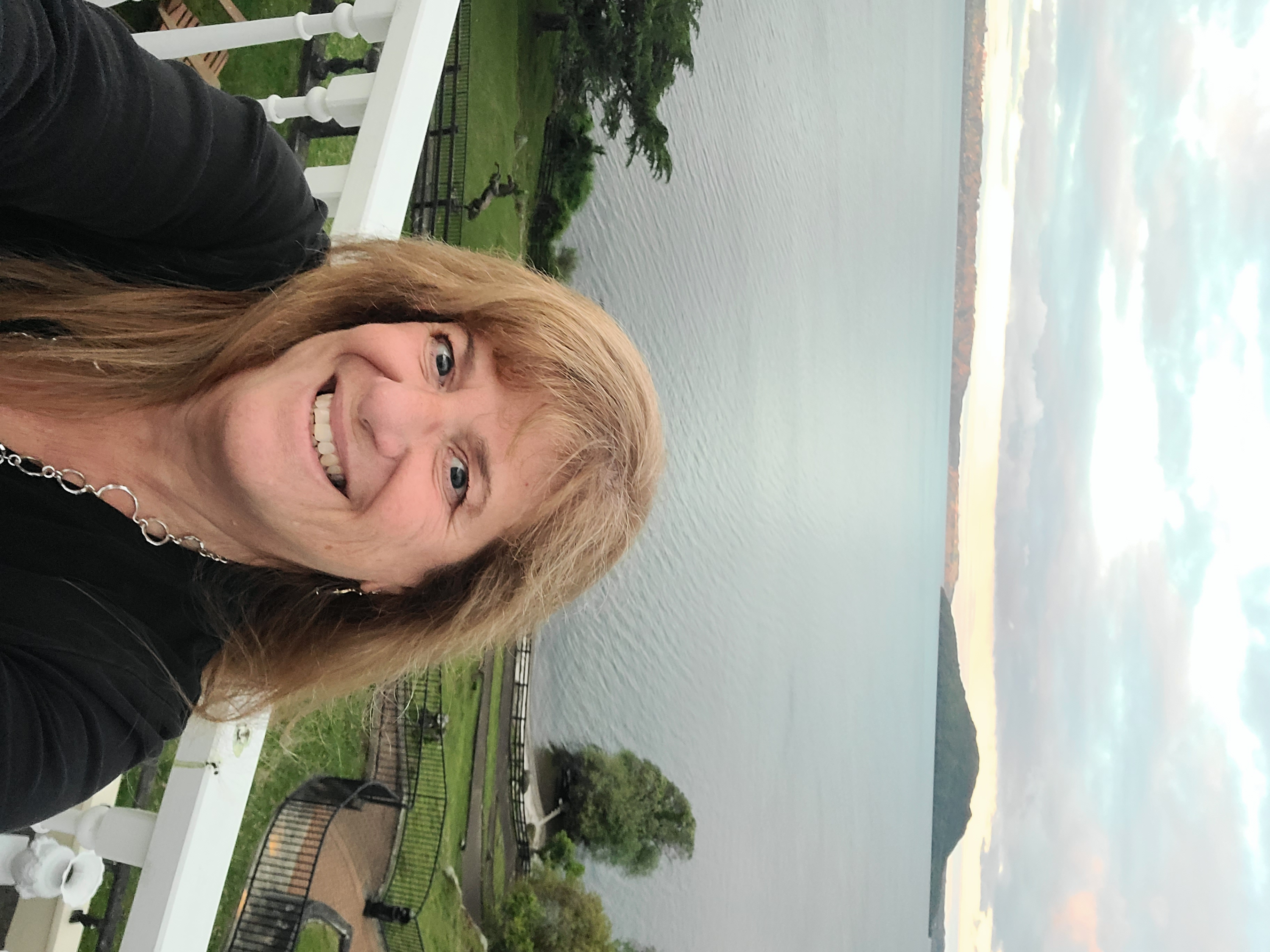Deborah smiling on a balcony with a lake, forested shores, and mountains visible in the background