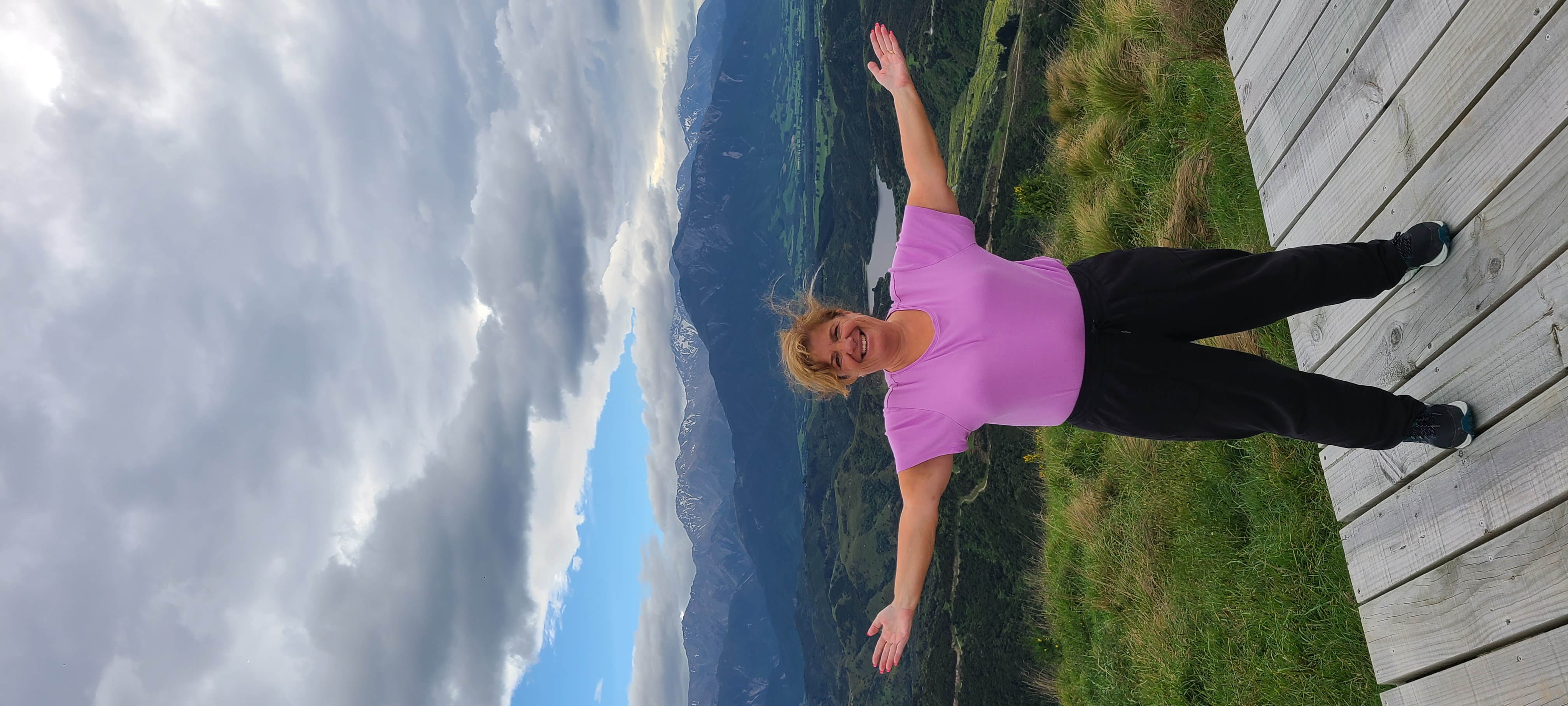 Deborah standing on wooden deck with arms raised, overlooking a lake and mountains in New Zealand