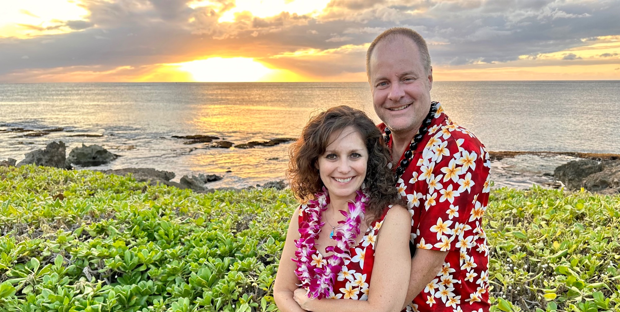 A man and woman wearing Hawaiian lei pose together at sunset on a rocky Hawaiian coastline with ocean waves behind them