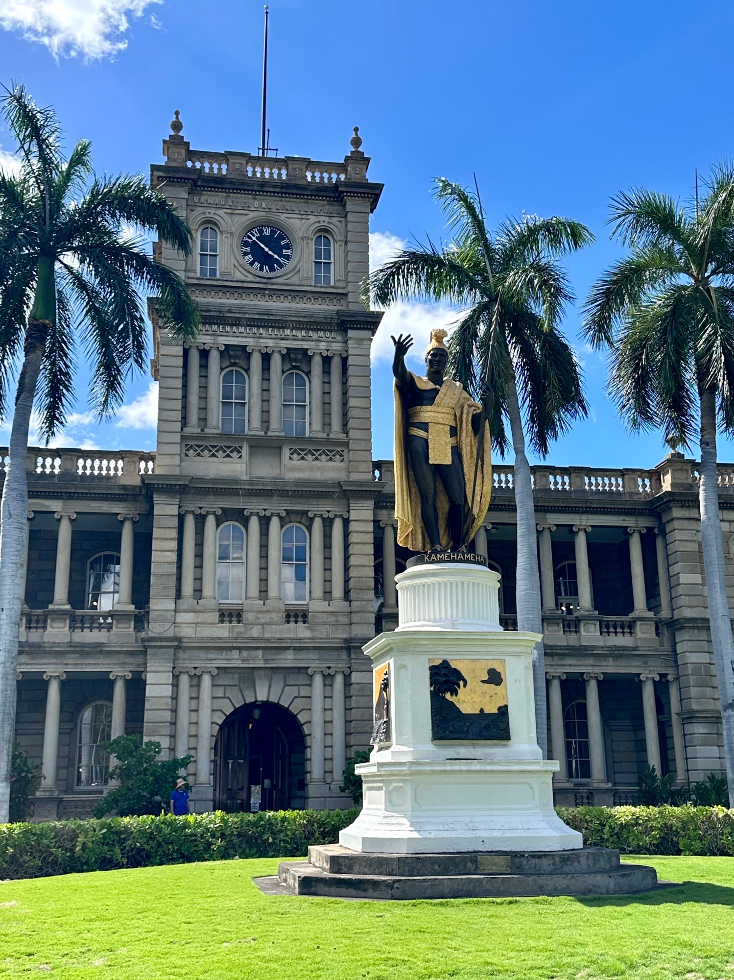 A golden statue of King Kamehameha I stands in front of a historic stone government building with a clock tower in Honolulu