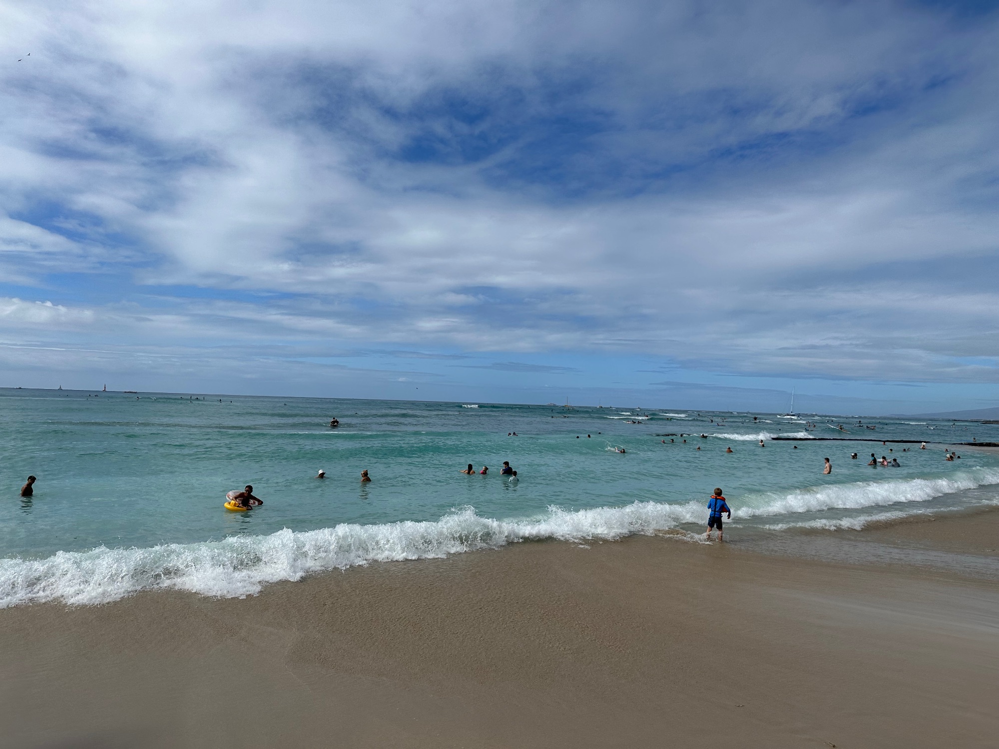 Beachgoers swim and play in turquoise water with white-capped waves at a sandy Hawaiian beach under blue skies