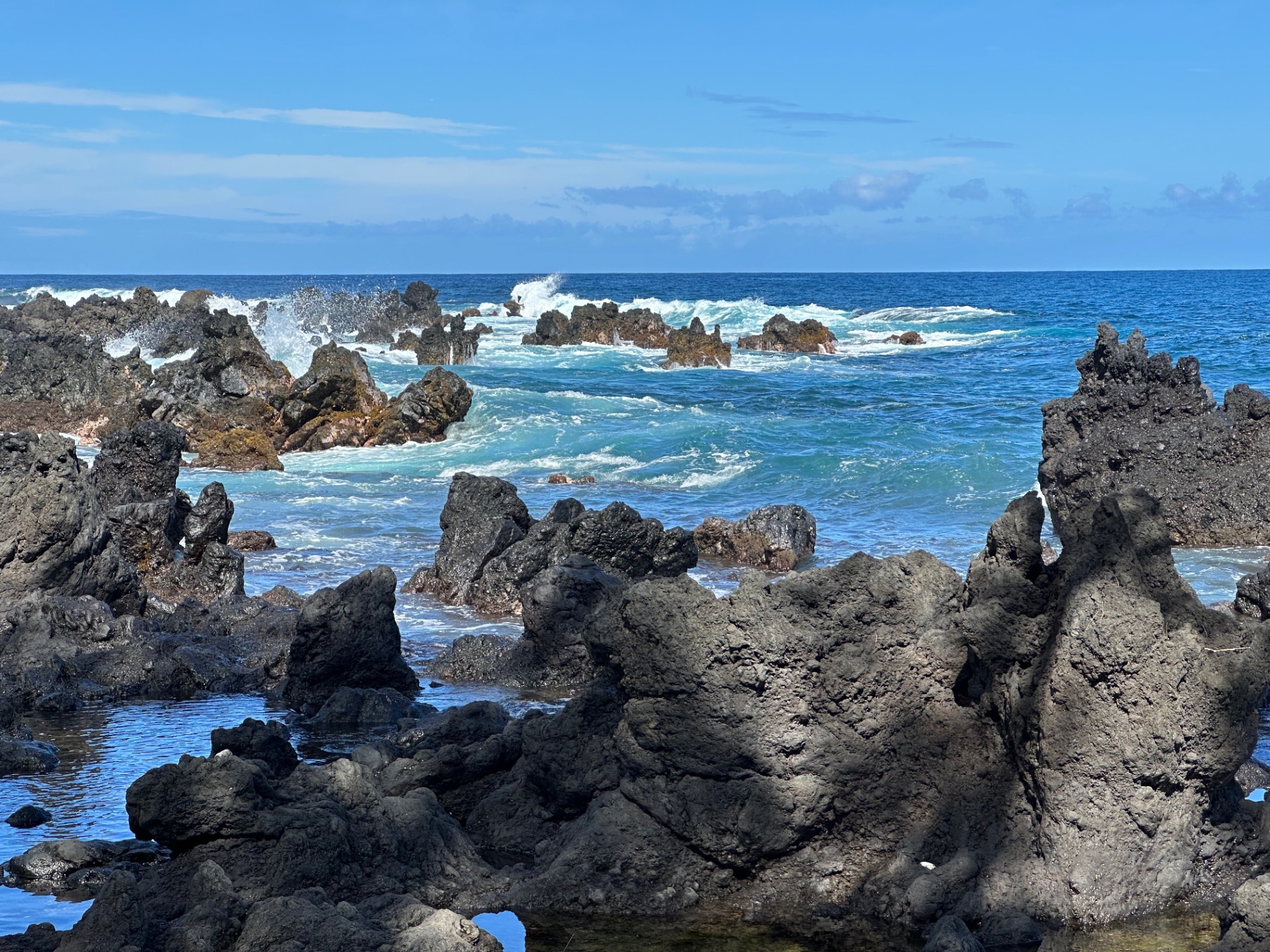 Dark volcanic rock formations jut from turquoise ocean water with white waves crashing against them on a Hawaiian coast