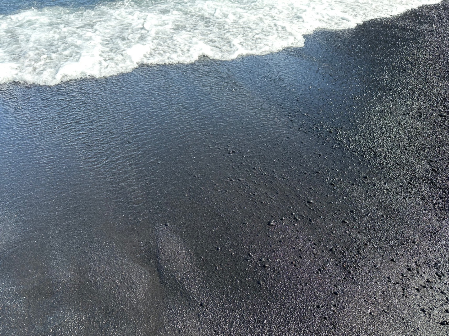 A black sand beach meets white foaming waves under a partly cloudy sky on a Hawaiian island