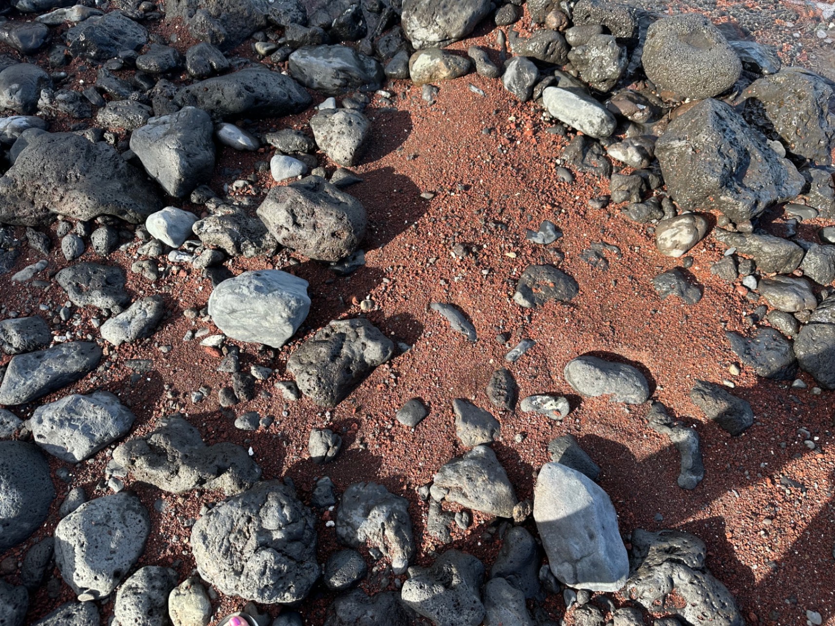 Reddish-brown volcanic rocks and pebbles scatter across a dark sand beach with white ocean waves in the background
