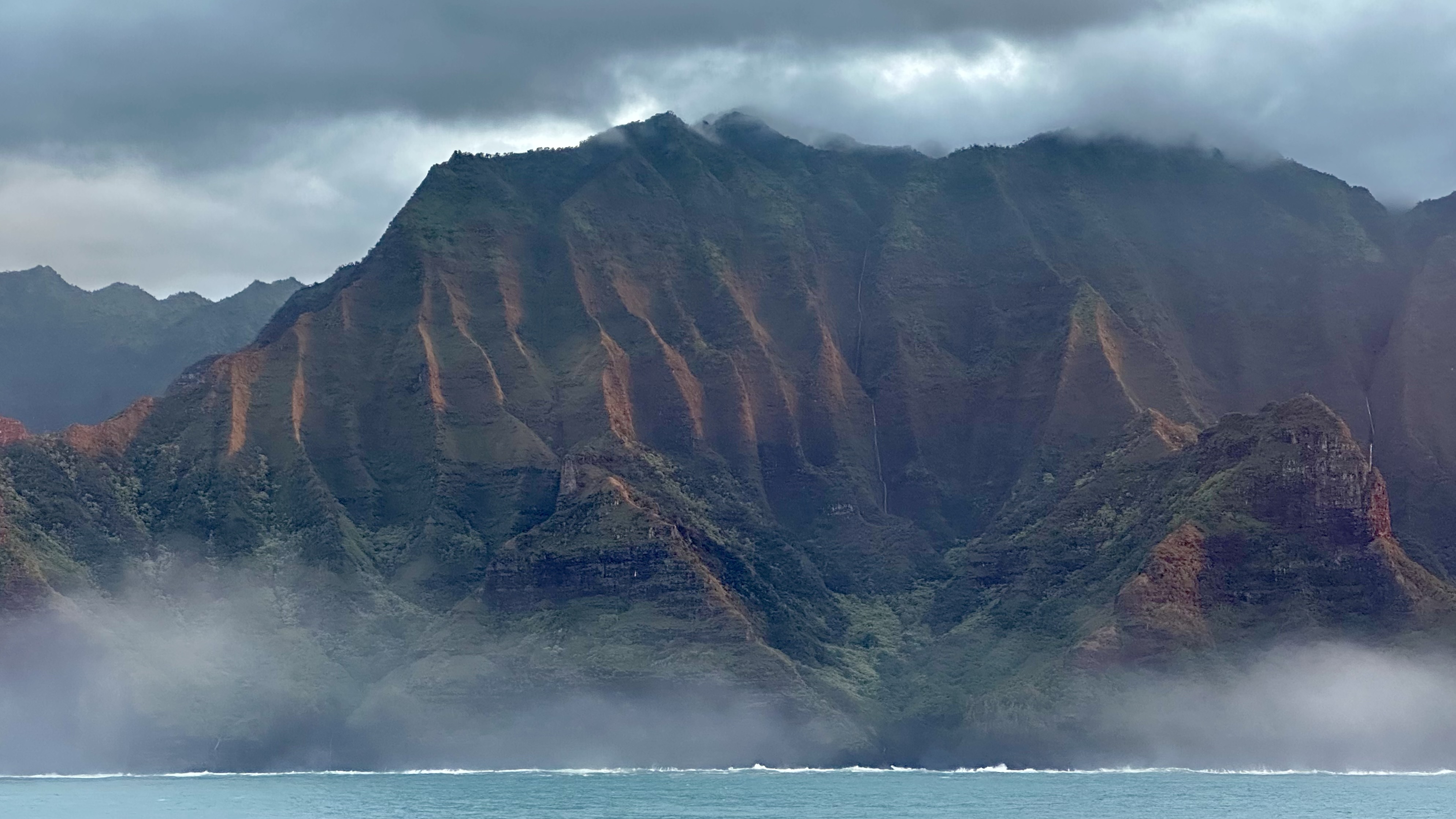 Dramatic sea cliffs with vertical ridges and waterfalls rise from turquoise ocean waters on the Hawaiian coast