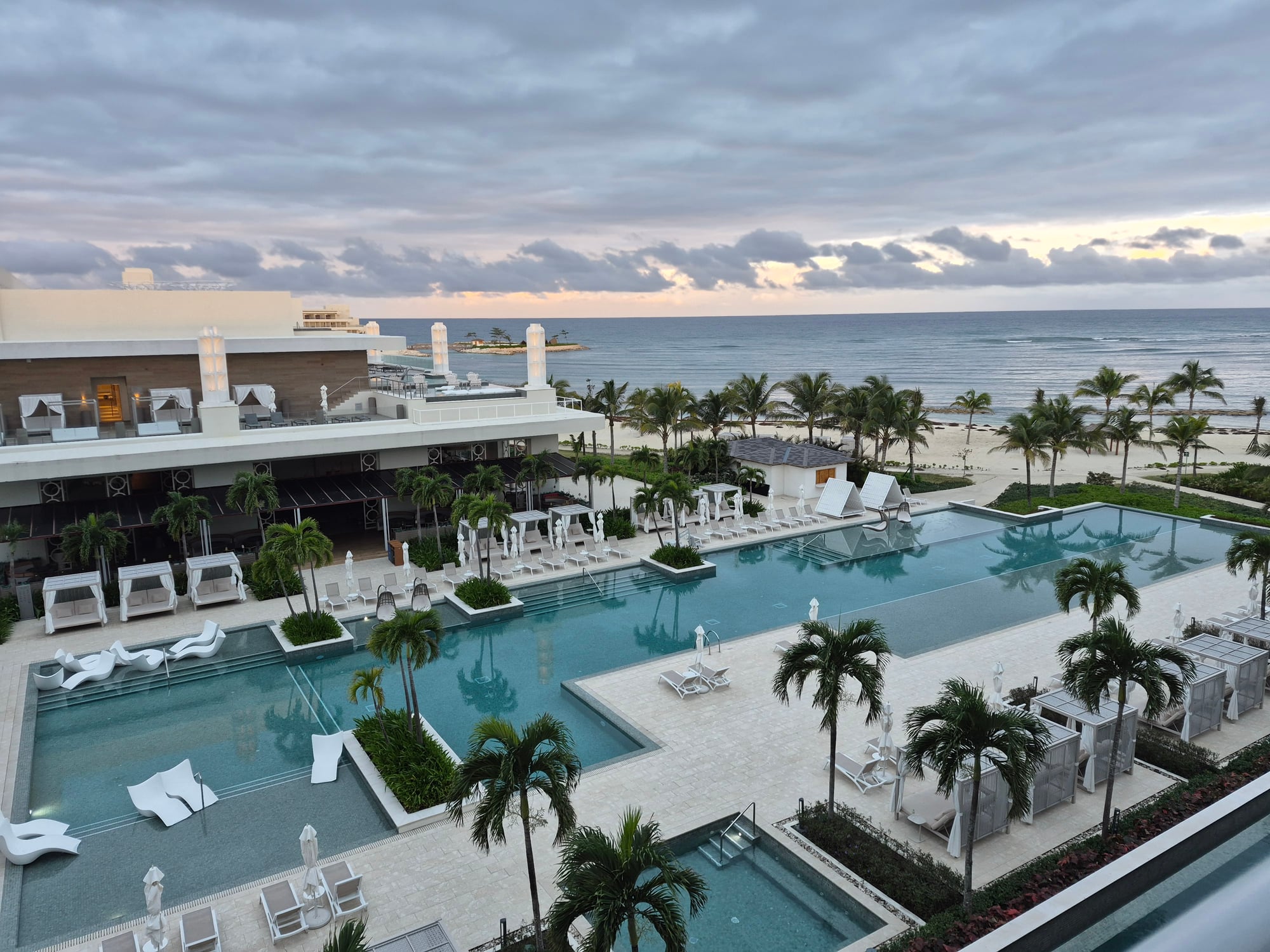 Resort pool area at dusk with illuminated turquoise water, white loungers, palm trees, and modern architecture overlooking the ocean