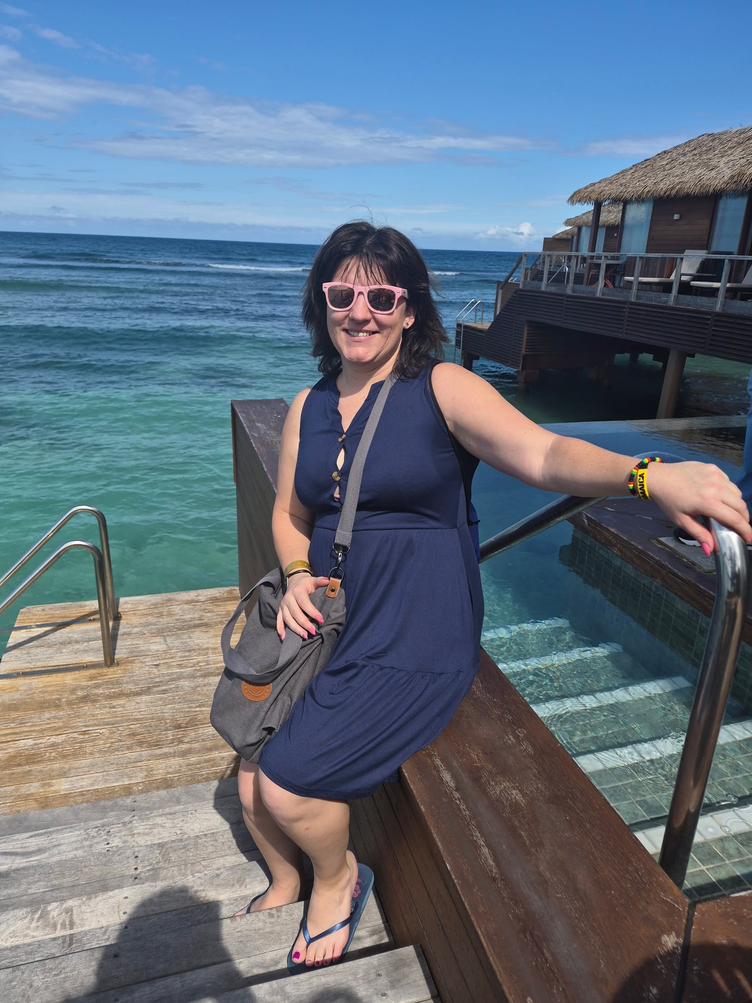 Woman in navy dress smiling on wooden dock with turquoise ocean and overwater bungalows in background
