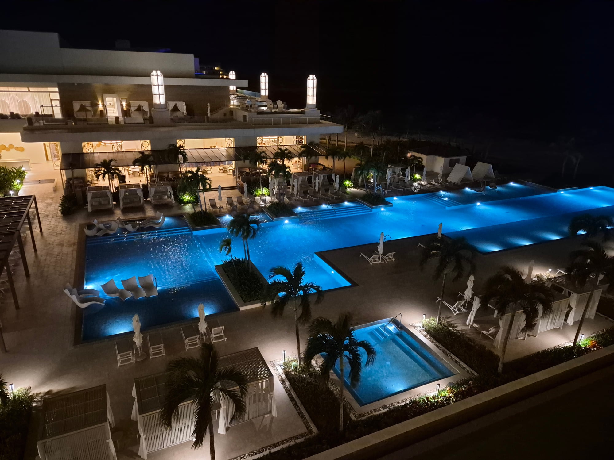 Nighttime aerial view of illuminated resort pools and grounds with white loungers and palm trees