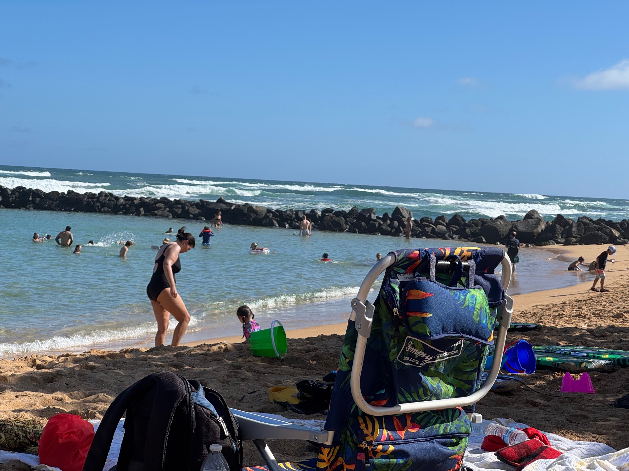 A beach scene with swimmers and families enjoying the water near dark volcanic rocks, with colorful beach gear and towels in the foreground