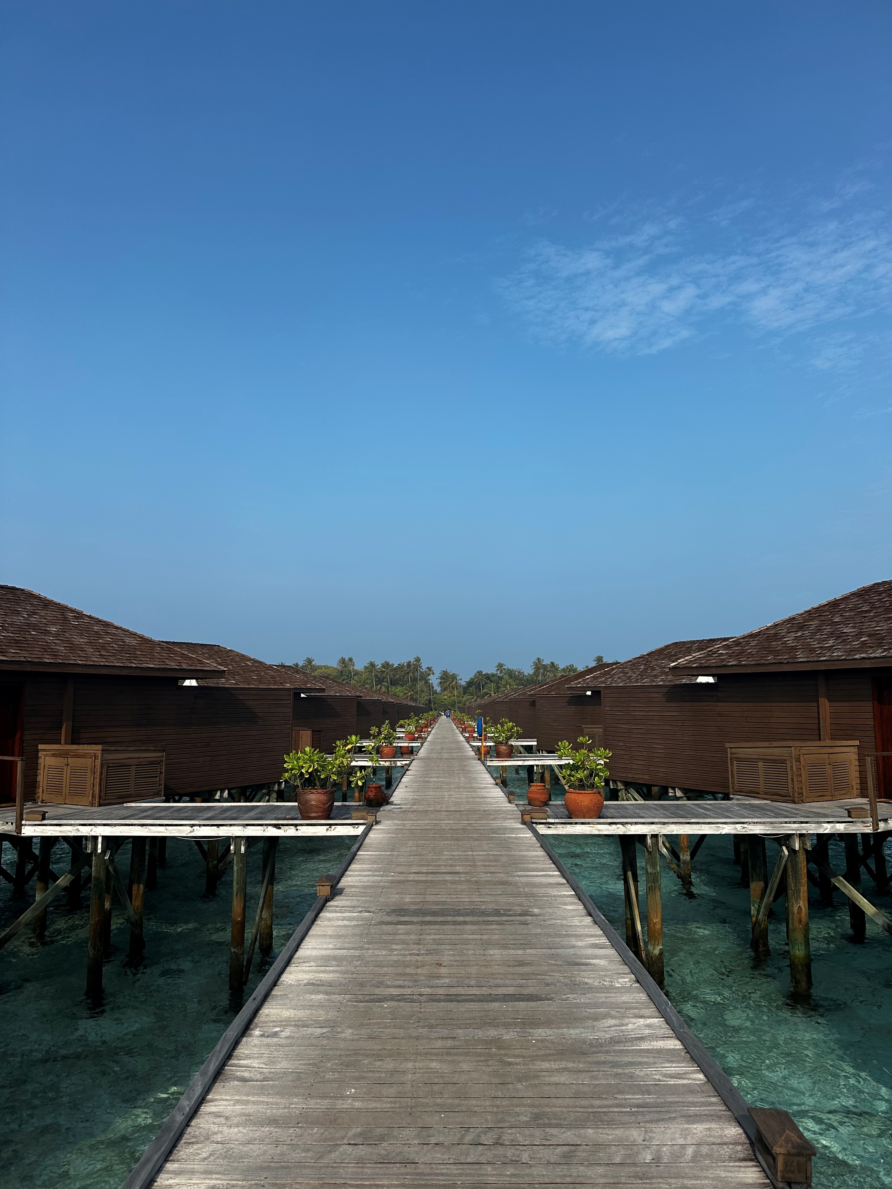 A wooden boardwalk stretches toward overwater bungalows with dark roofs on either side under clear blue sky