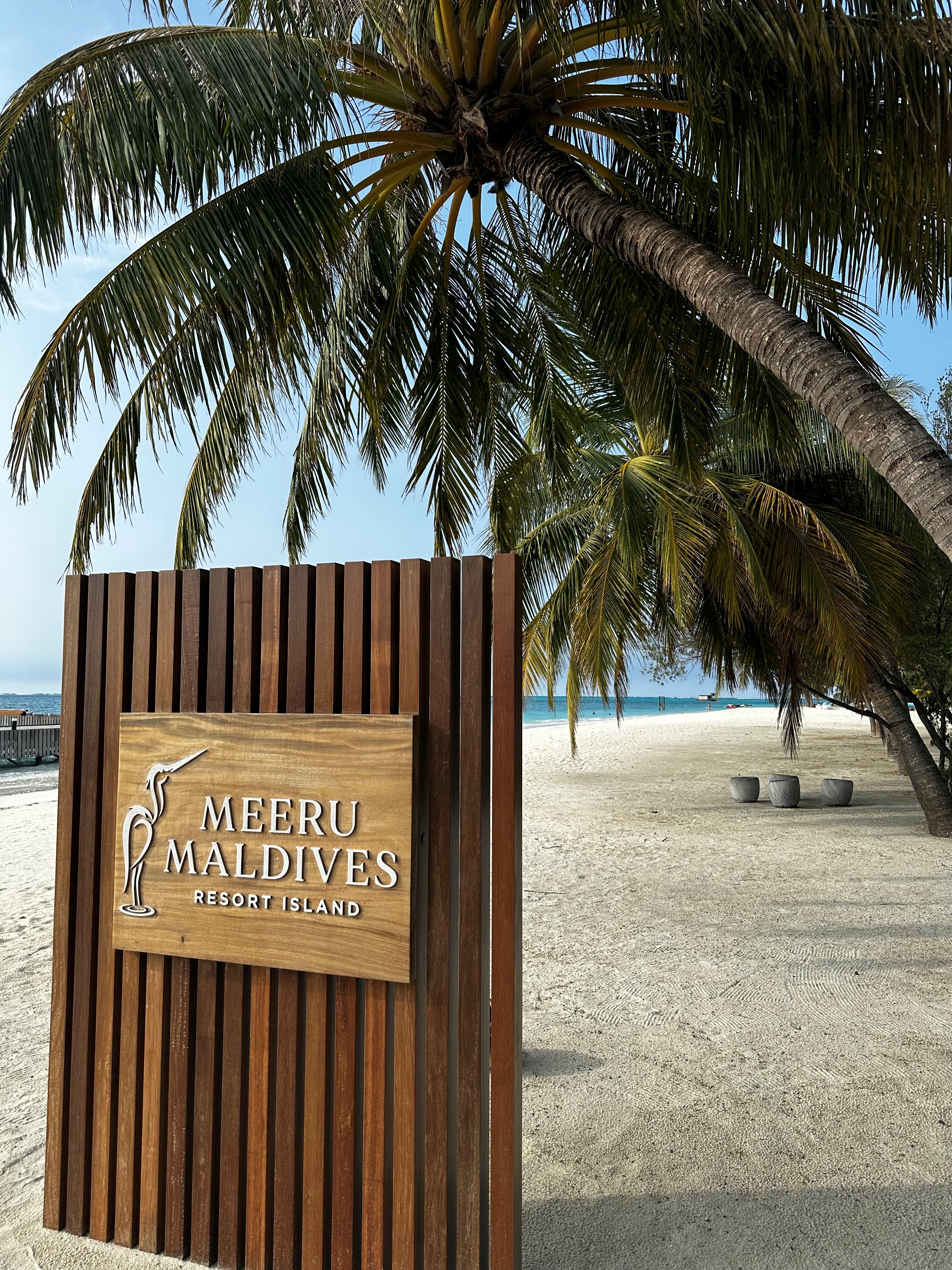 Meeru Maldives Resort Island wooden sign on a beach with palm tree and white sand visible