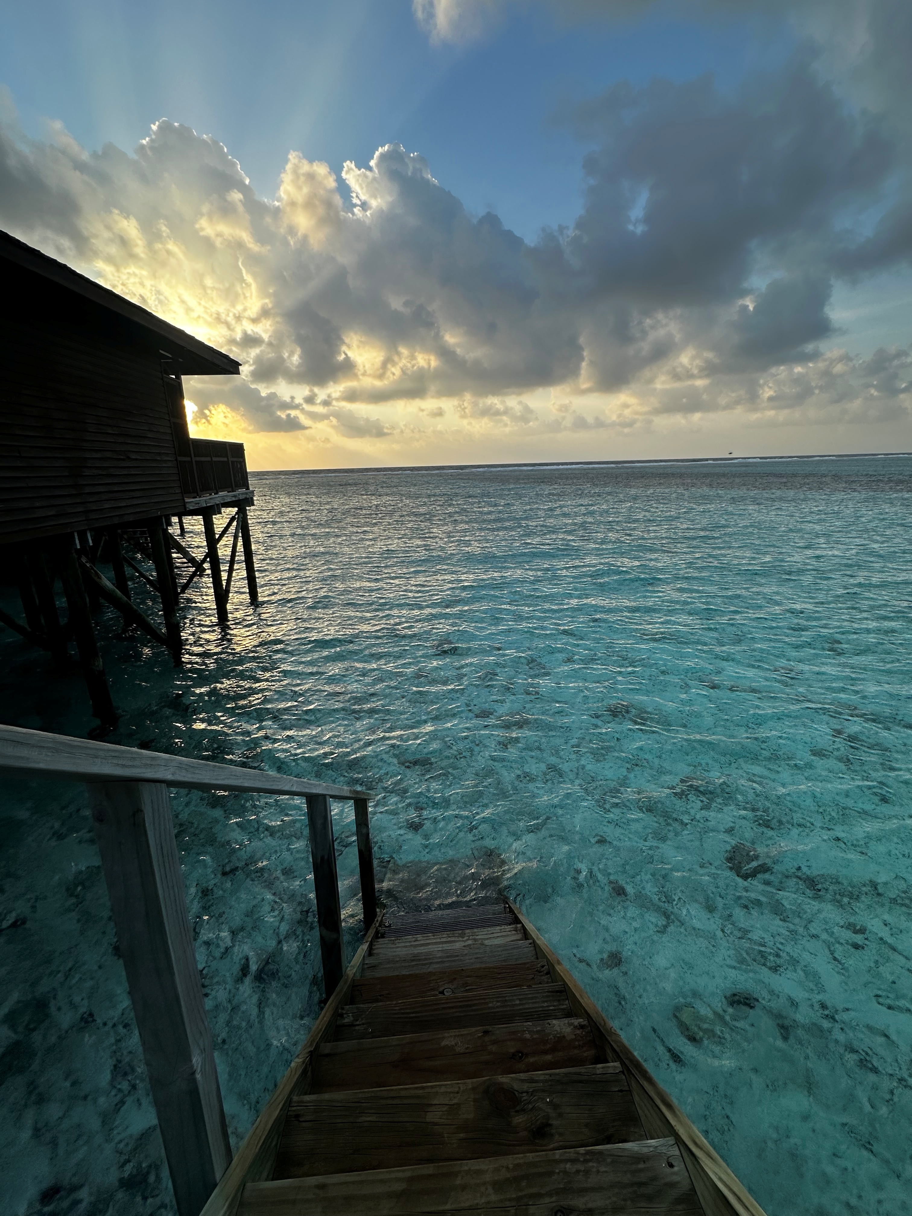 Sunset view from an overwater bungalow deck with dramatic clouds illuminated in gold and turquoise ocean below