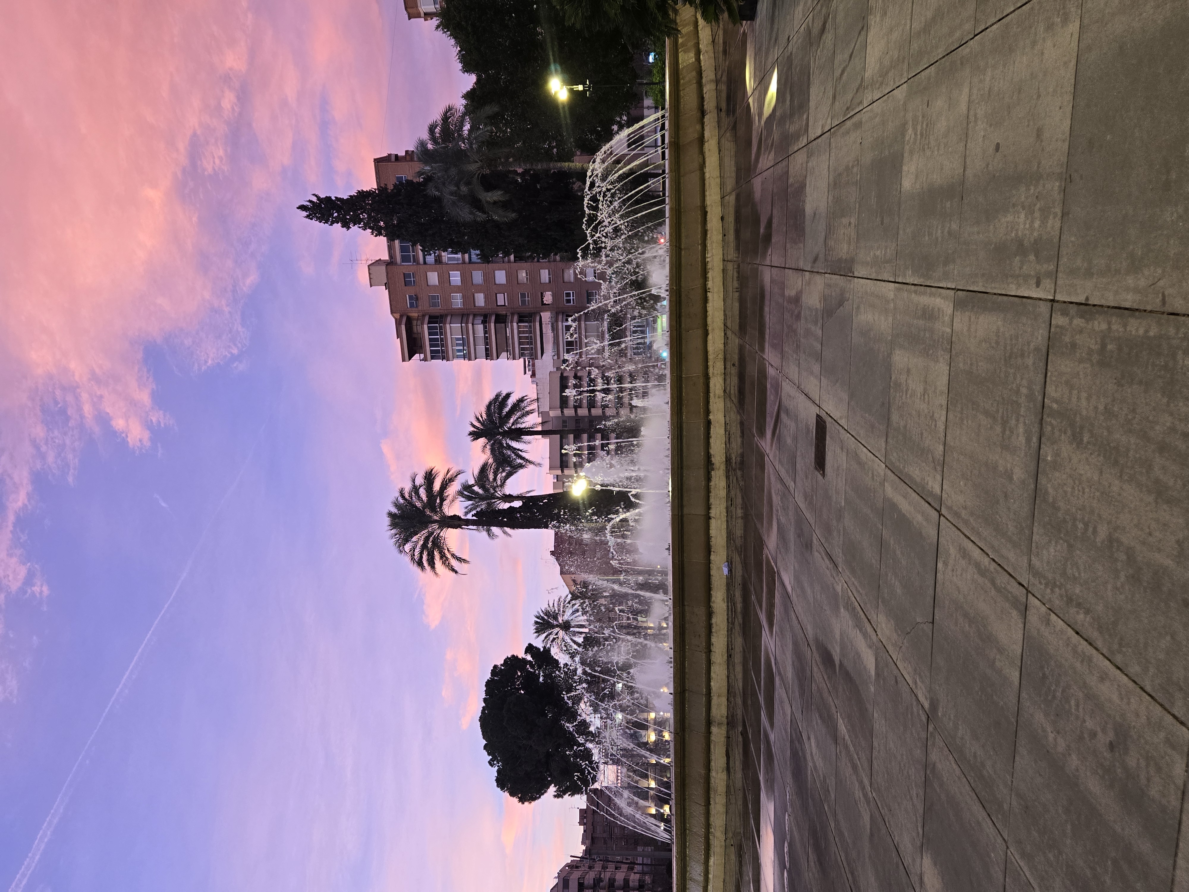 A fountain with illuminated water jets at dusk in a Madrid plaza with palm trees