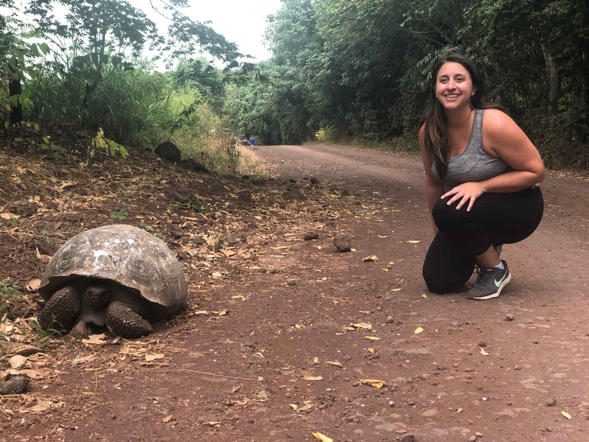 Beth Sharry crouching next to a Galápagos tortoise on a dirt trail surrounded by vegetation