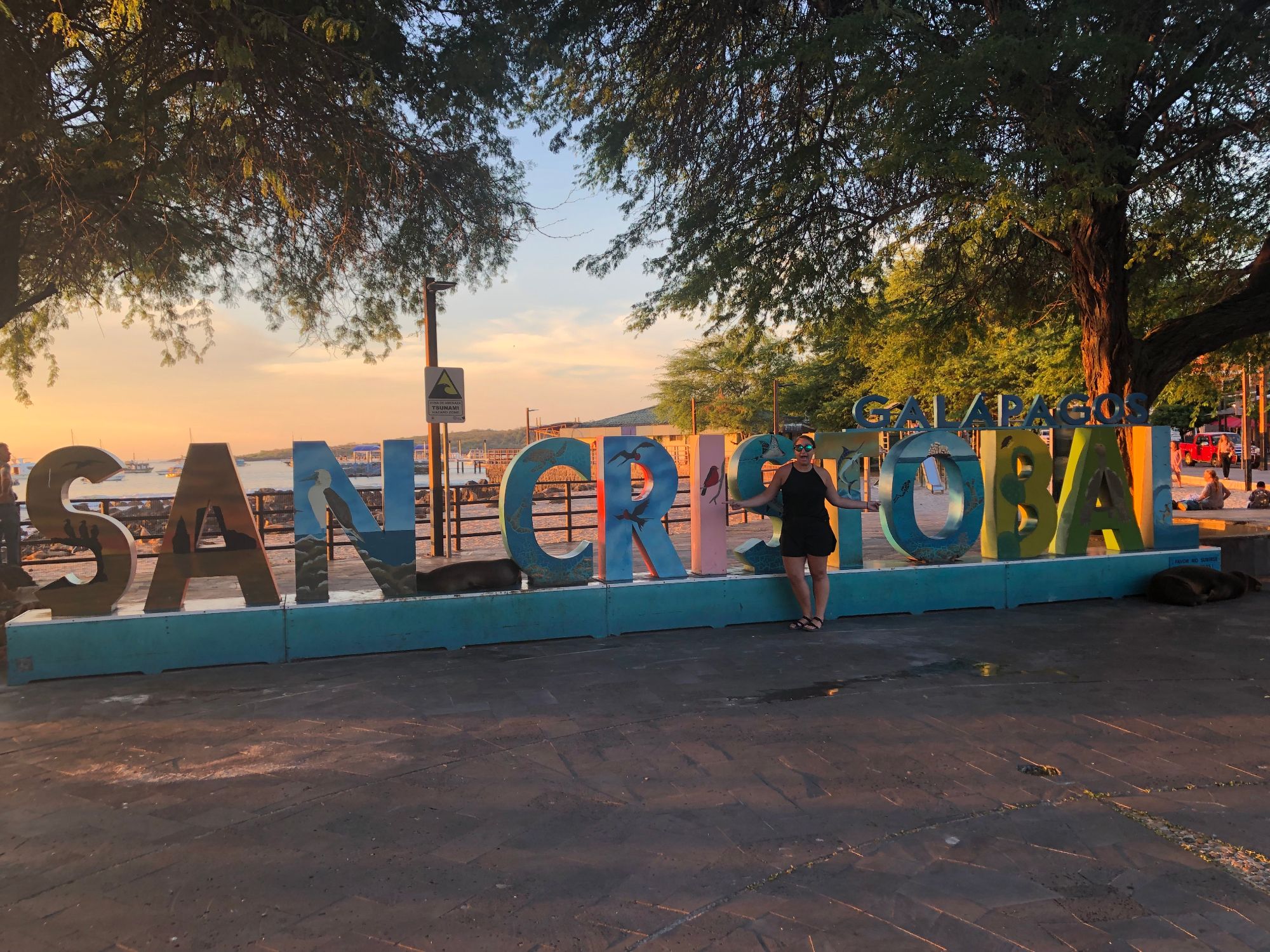 Beth Sharry at the San Cristóbal waterfront sign at sunset with boats and ocean visible