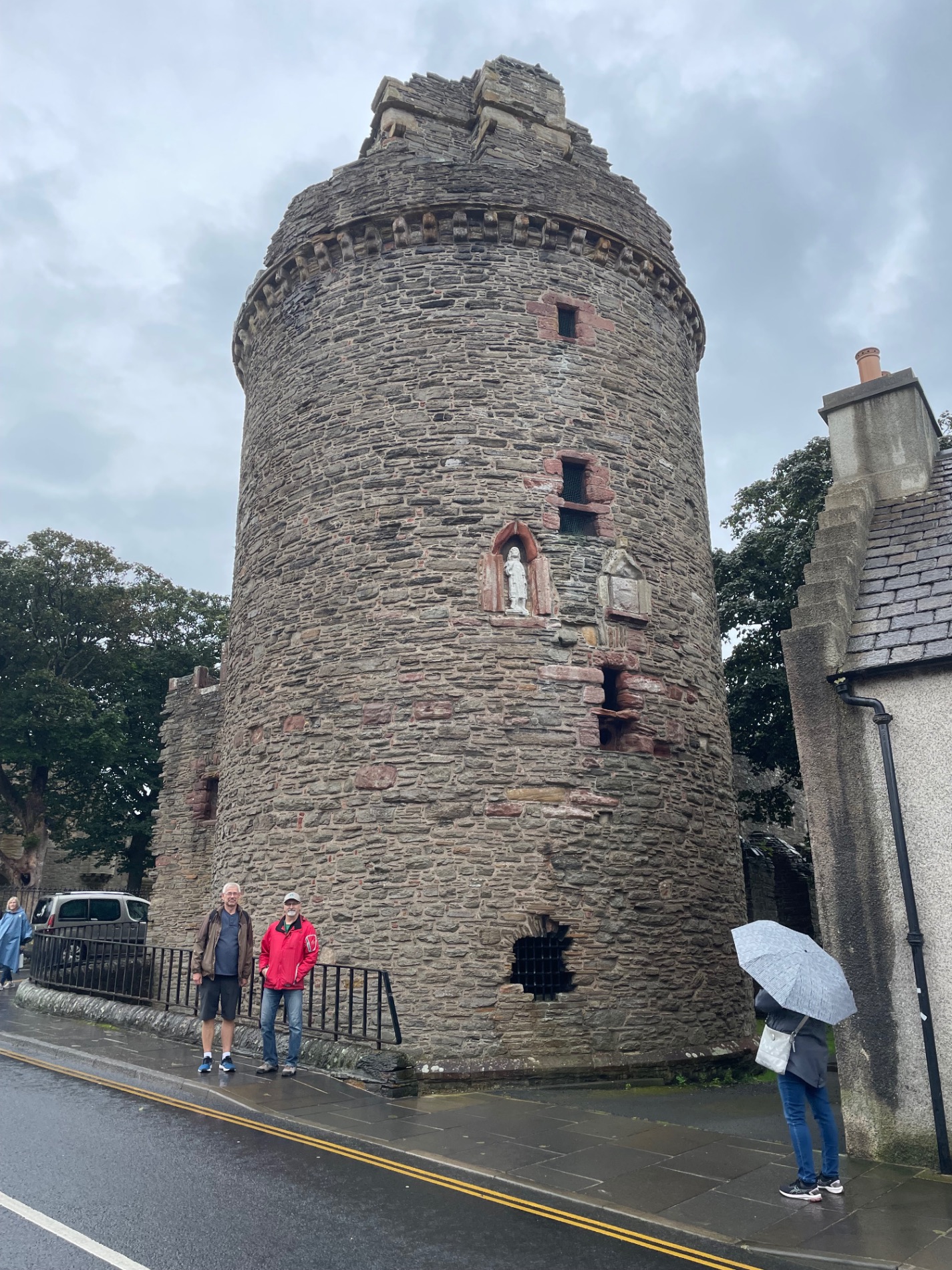 Two visitors stand near a historic stone tower in Scotland on a rainy day, one holding an umbrella