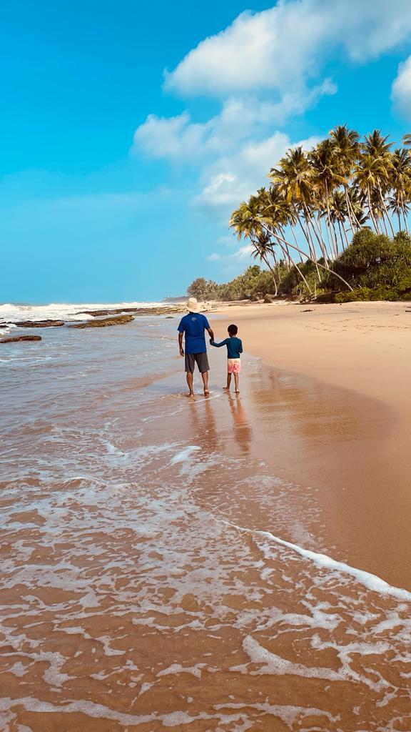 Adult and child wading together in shallow ocean water on a sandy Sri Lankan beach lined with palm trees