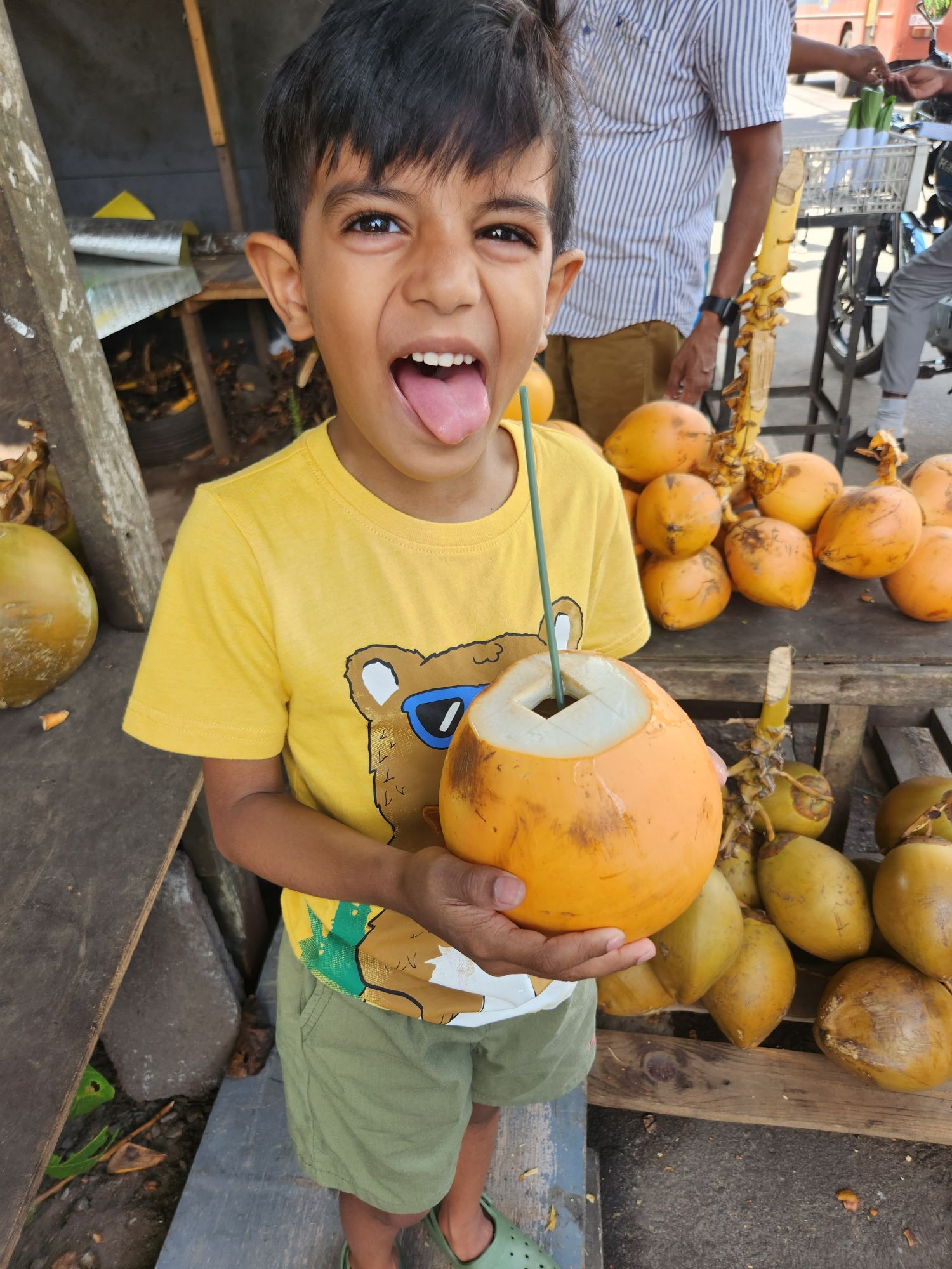 Young boy in yellow shirt holding a fresh coconut with a straw at a Sri Lankan market stall