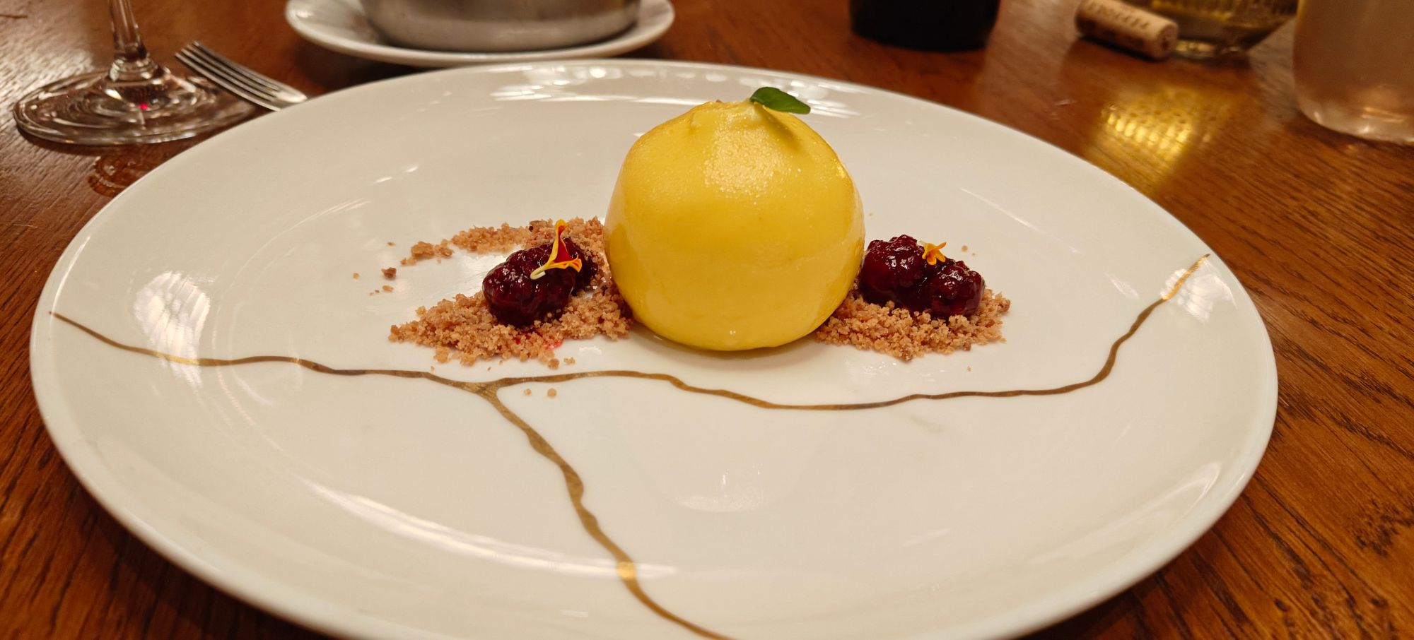 Plated dessert with a yellow sphere surrounded by crumble, dark berries, and decorative sauce lines on white plate