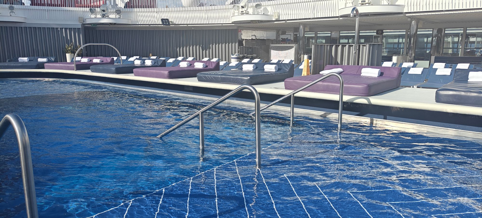 Resort pool deck with multiple lounge chairs in purple and black, metal railings, and covered seating areas under bright sunlight