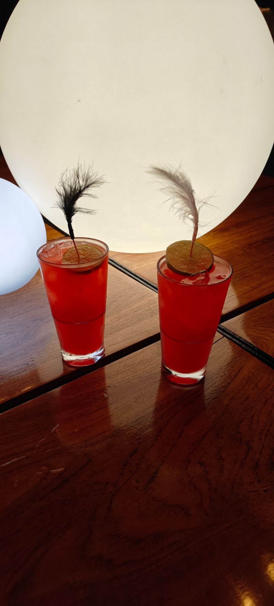Two red cocktails garnished with lime wheels and decorative grass sprigs on wooden table with white globe lamp