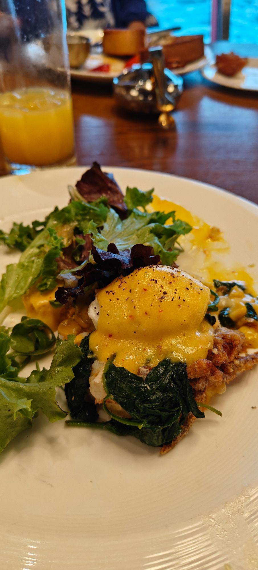 Plated egg Benedict with spinach, crispy base, and mixed greens salad with orange juice in background at dining table